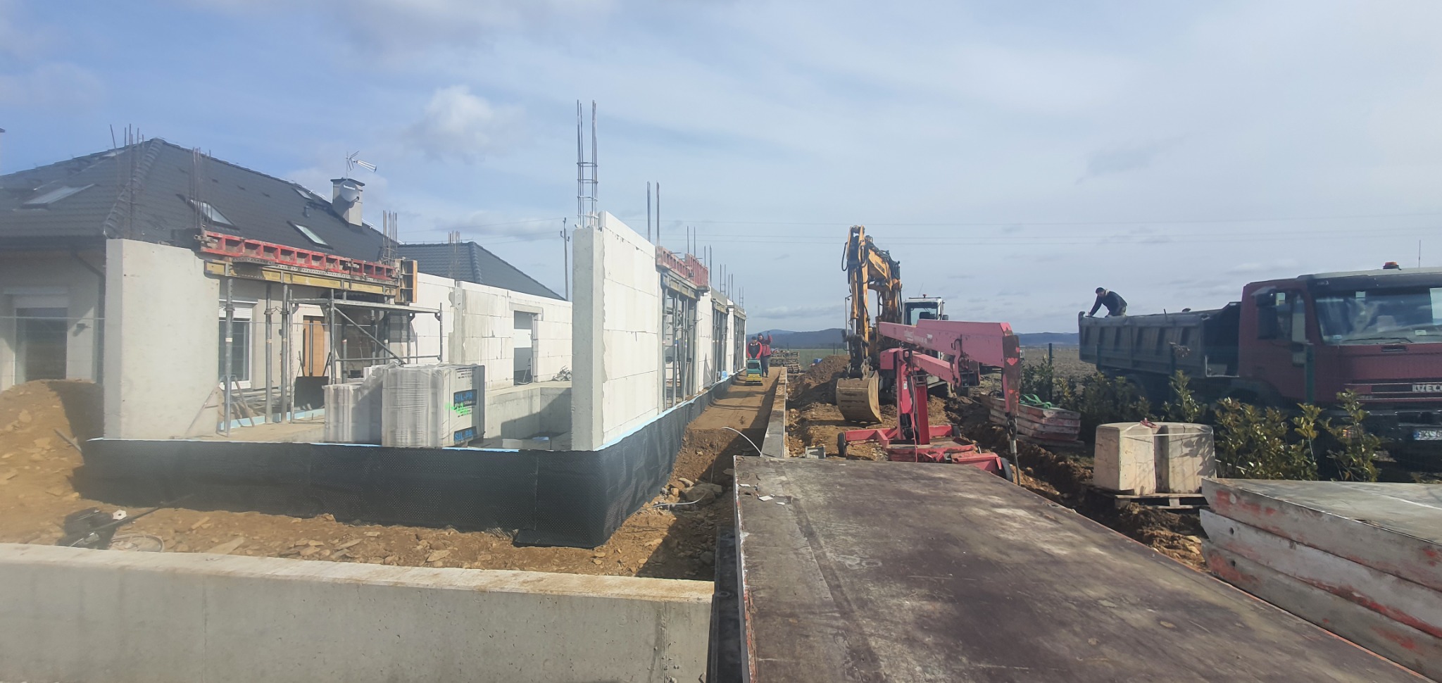 Budowa domu jednorodzinnego w trakcie wznoszenia ścian z bloczków, widoczna izolacja fundamentów, sprzęt budowlany w tle oraz pracownicy na terenie budowy.