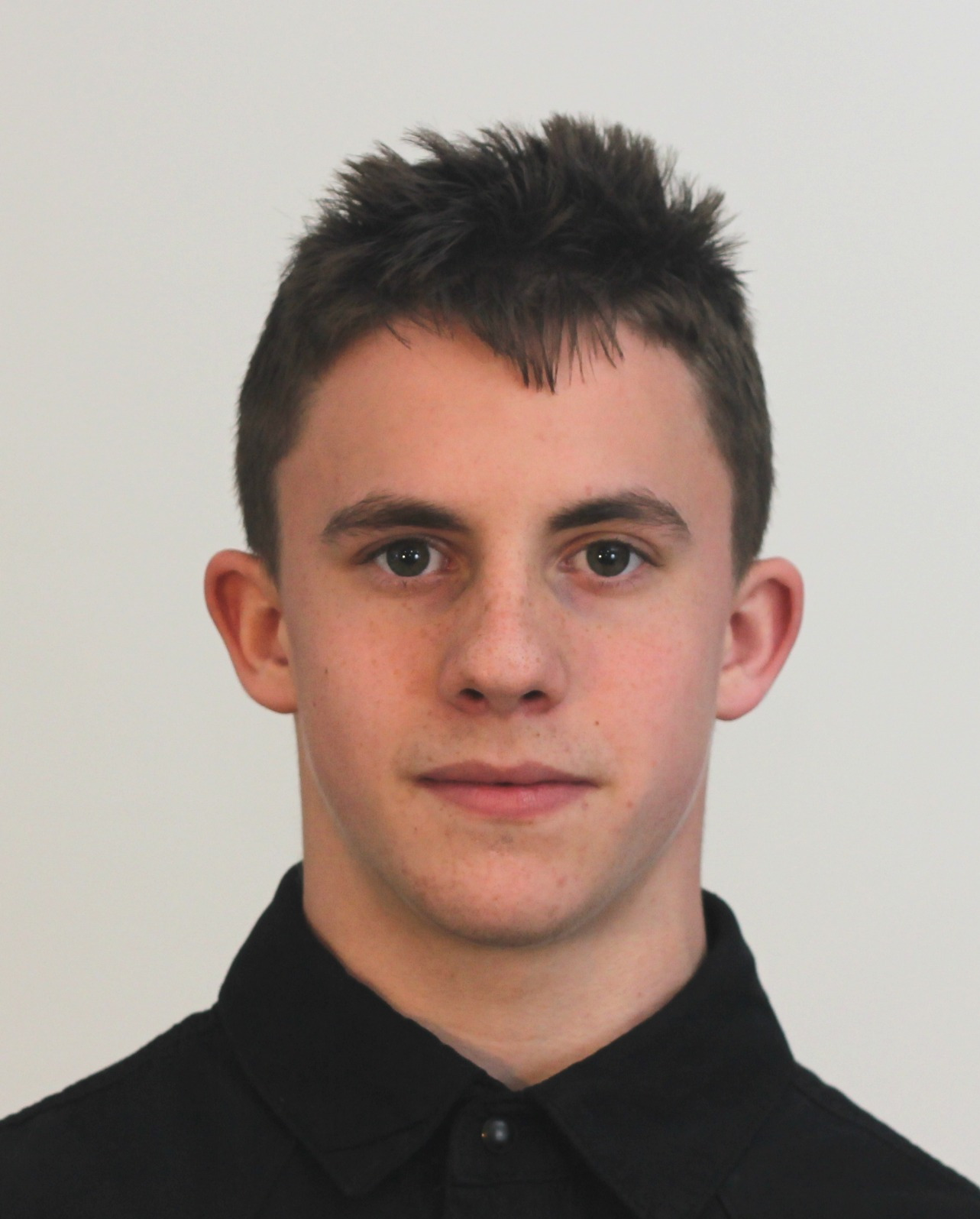 Portret młodego mężczyzny z krótkimi, ciemnymi włosami i ciemną koszulą na jasnym tle, wykonany w neutralnym oświetleniu.