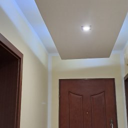 Firma uslugowa - Pomieszczenie po remoncie: drewniane drzwi, kremowe ściany i sufit z podwieszaną konstrukcją oraz oświetleniem LED. Widok z dołu, skupienie na suficie.