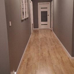 Firma uslugowa - Długi korytarz po malowaniu ścian na szaro, z drewnianą podłogą. Widoczne puszki z farbą i taśma malarska. Na ścianie wiszą obrazy w białych ramach.