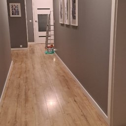 Firma uslugowa - Szary korytarz po malowaniu, z panelami podłogowymi, białymi listwami przypodłogowymi i drabiną malarską w tle. Widoczne trzy obrazy w białych ramach.