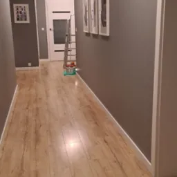 Szary korytarz po malowaniu, z panelami podłogowymi, białymi listwami przypodłogowymi i drabiną malarską w tle. Widoczne trzy obrazy w białych ramach.