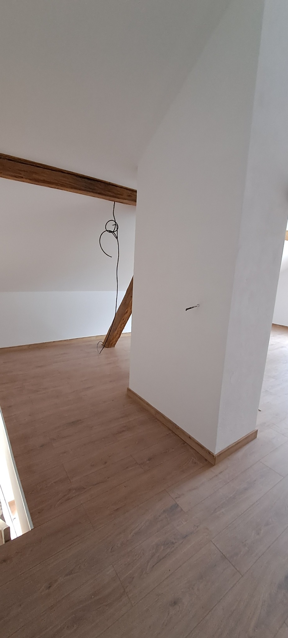 Wnętrze poddasza po remoncie: drewniana podłoga, białe ściany i drewniane belki stropowe. Widoczne wykończenia podłogowe przy ścianach i kolumnie.