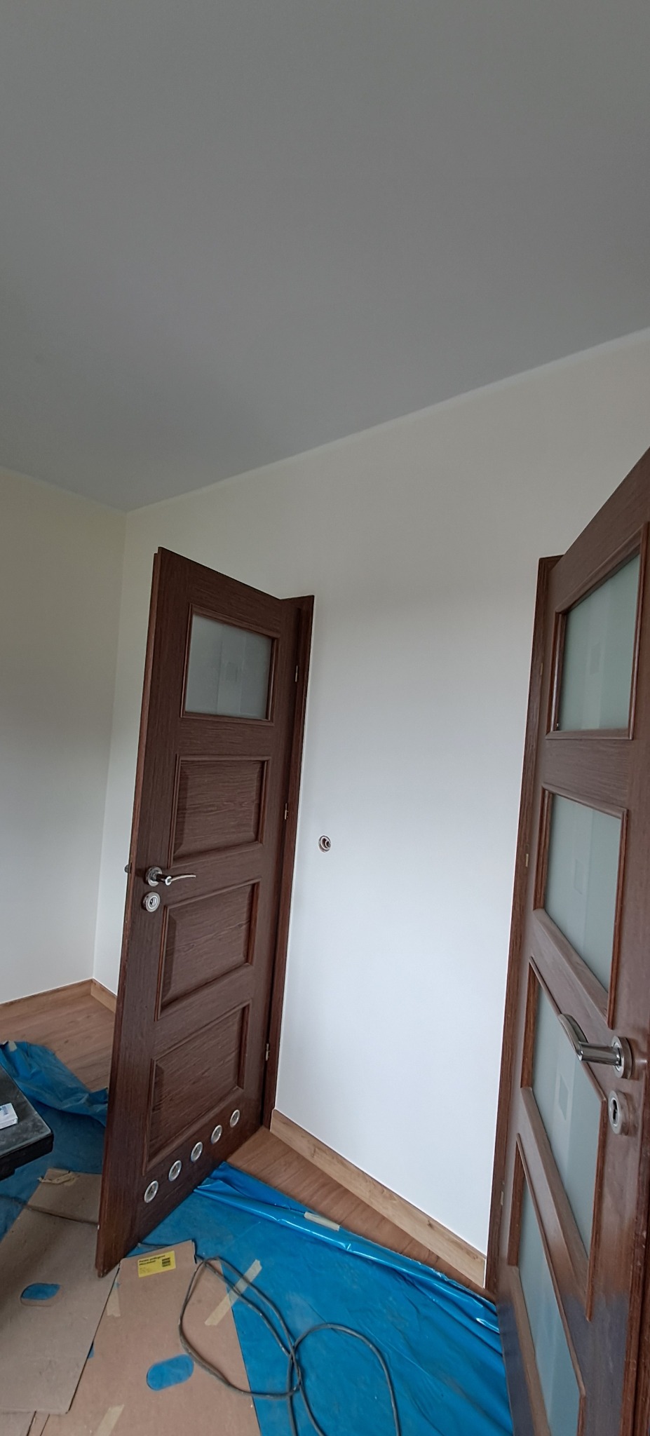 Dwa drewniane, przeszklone skrzydła drzwiowe po montażu w nowo wykończonym wnętrzu. Podłoga zabezpieczona folią malarską. Widoczne listwy przypodłogowe.