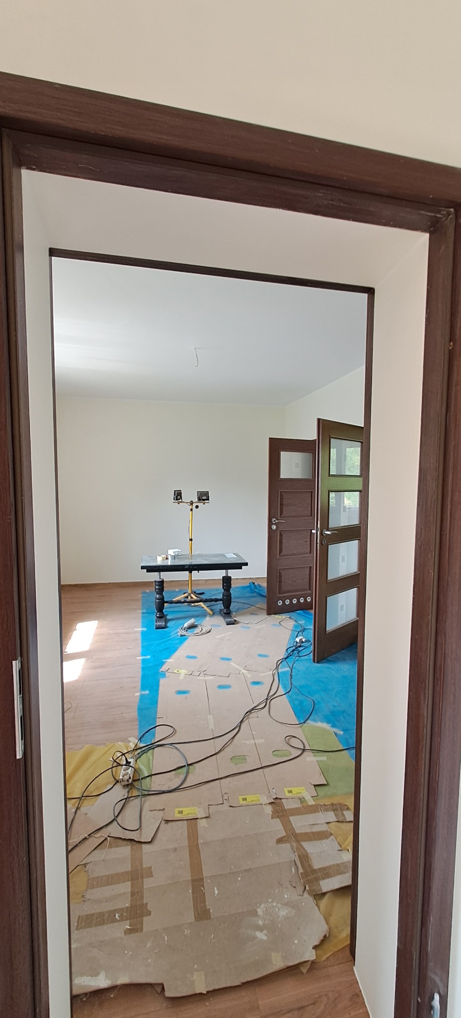 Widok przez futrynę na zabezpieczoną podłogę, stół z lampami i otwarte, brązowe drzwi z przeszkleniami. Przygotowanie do malowania.