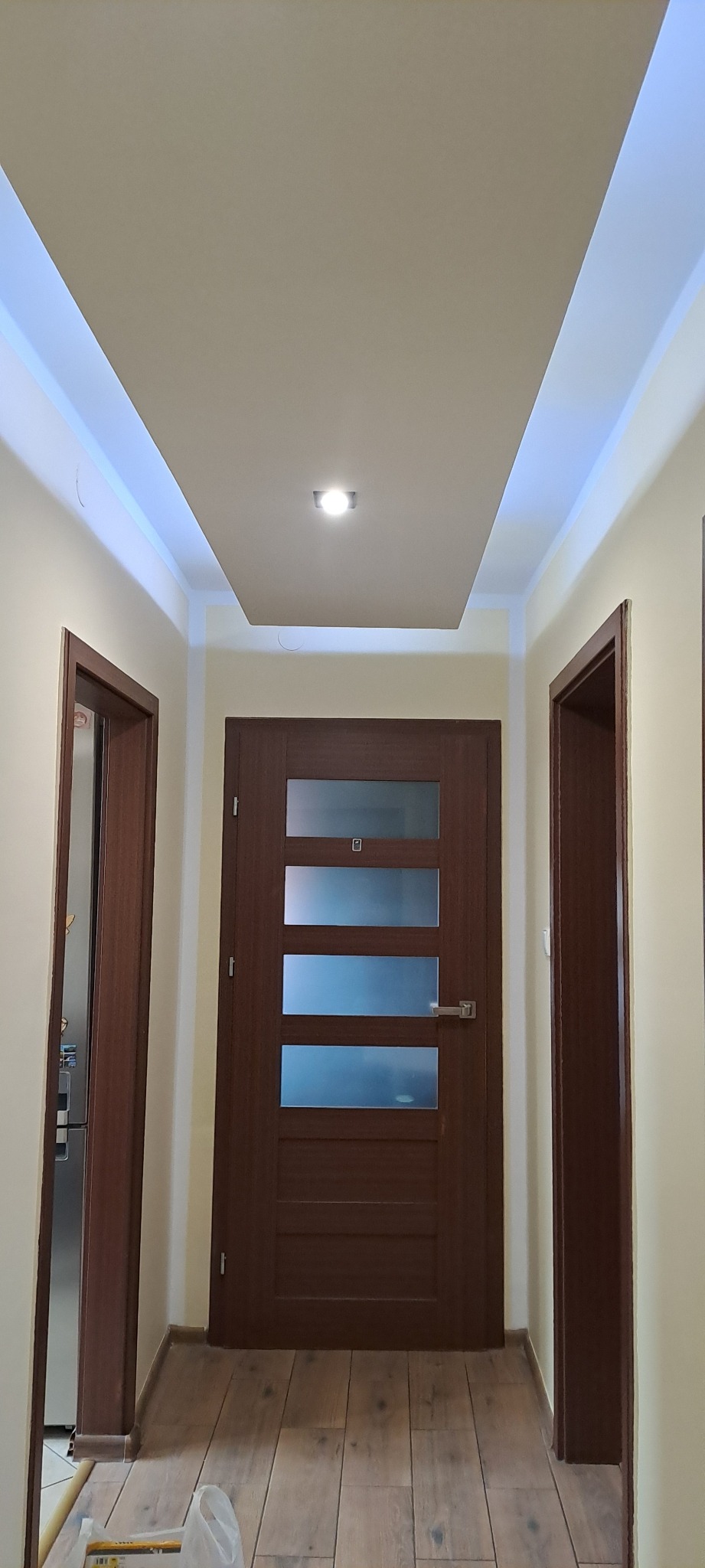 Korytarz po remoncie: drewnopodobna podłoga, brązowe drzwi z przeszkleniami i podwieszany sufit z oświetleniem LED. Widok na dwie pary drzwi.
