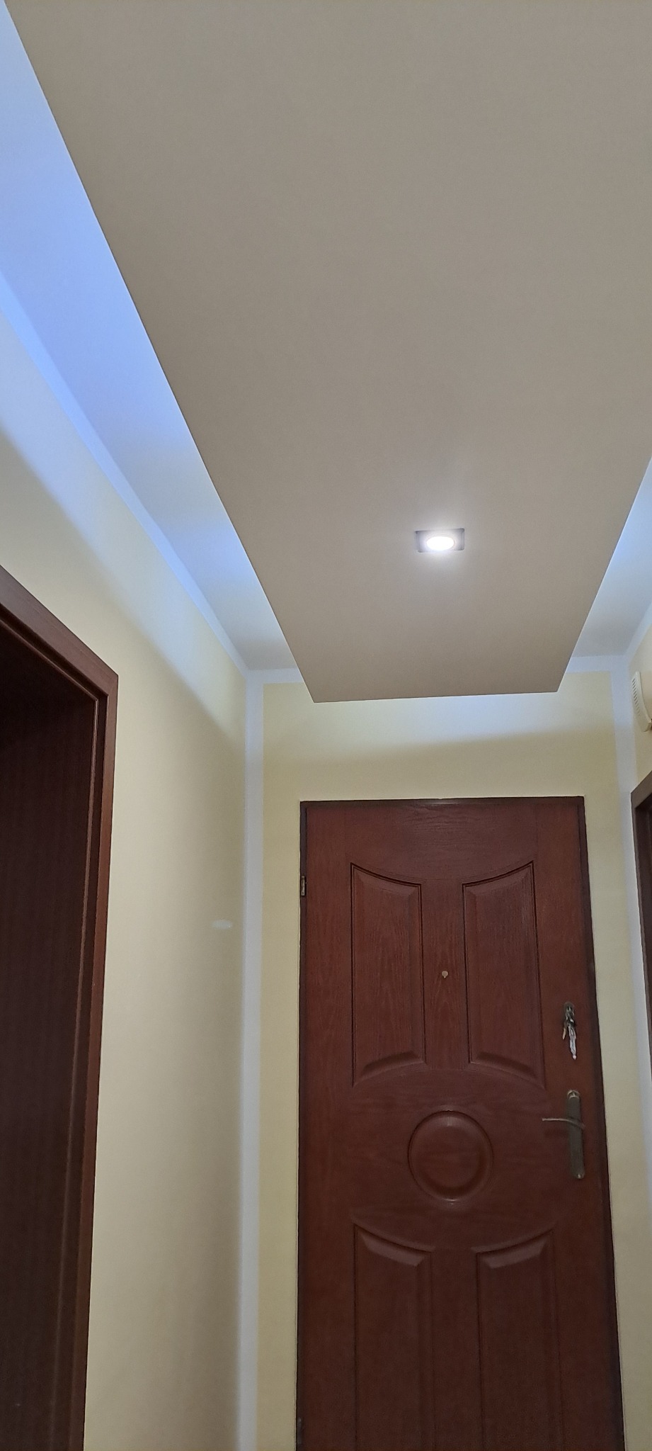 Pomieszczenie po remoncie: drewniane drzwi, kremowe ściany i sufit z podwieszaną konstrukcją oraz oświetleniem LED. Widok z dołu, skupienie na suficie.