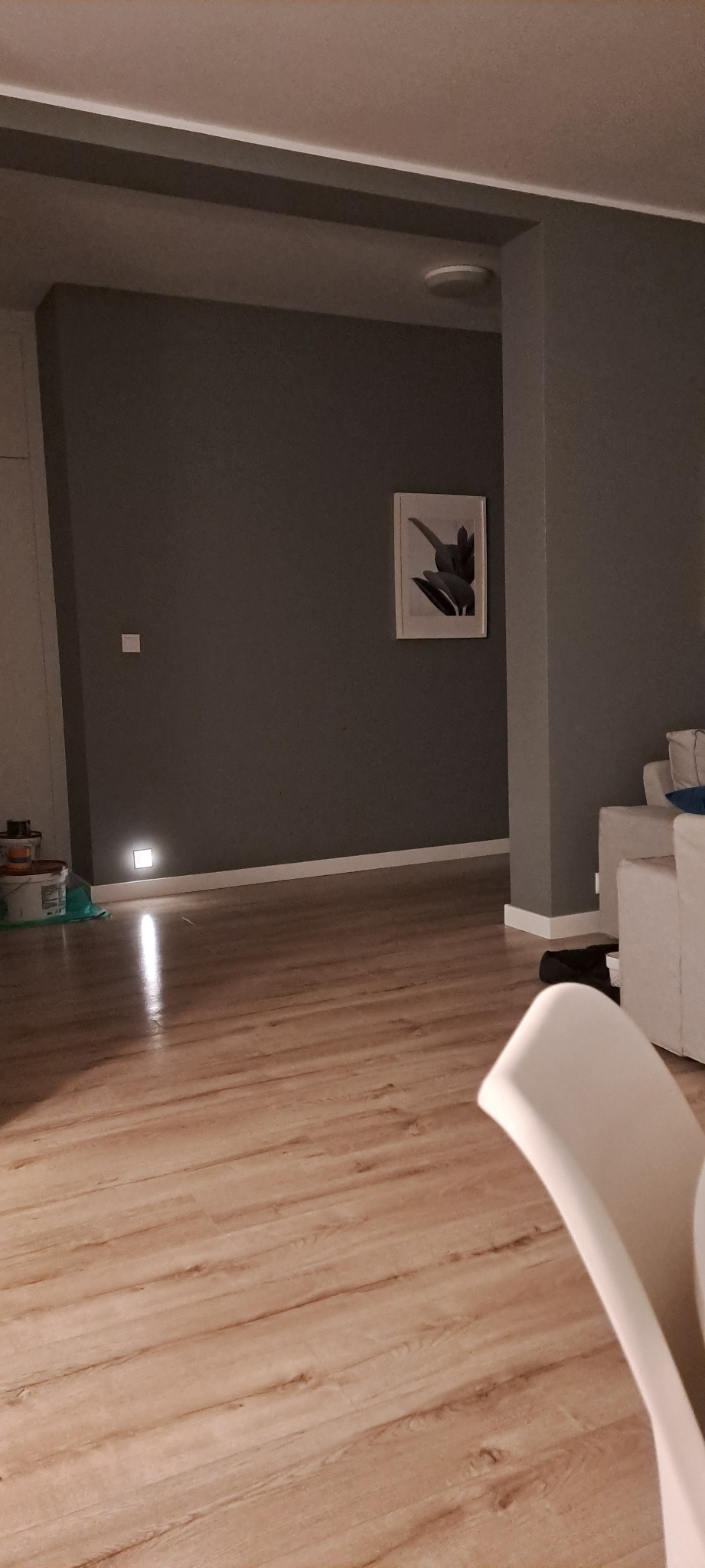 Szare ściany w pokoju z drewnianą podłogą, białą listwą przypodłogową i obrazem na ścianie. W tle widoczny biały narożnik i fragment krzesła.