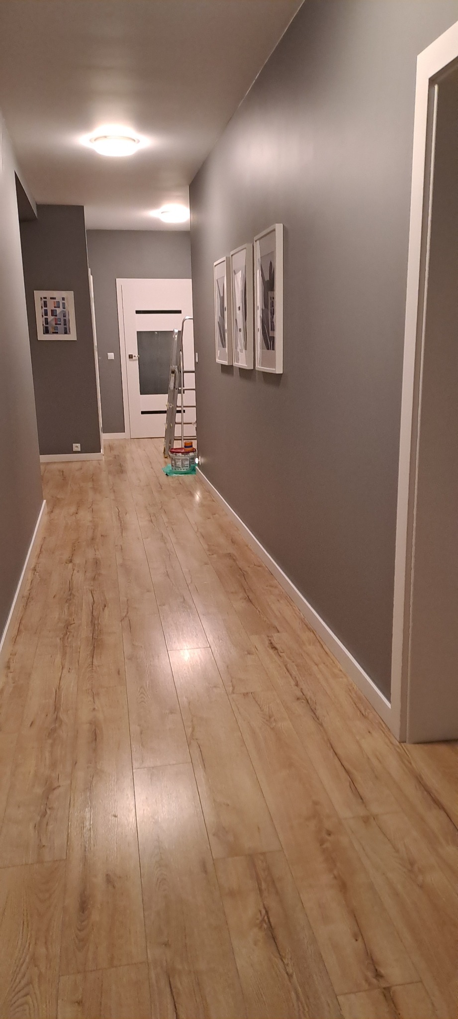 Szary korytarz po malowaniu, z panelami podłogowymi, białymi listwami przypodłogowymi i drabiną malarską w tle. Widoczne trzy obrazy w białych ramach.