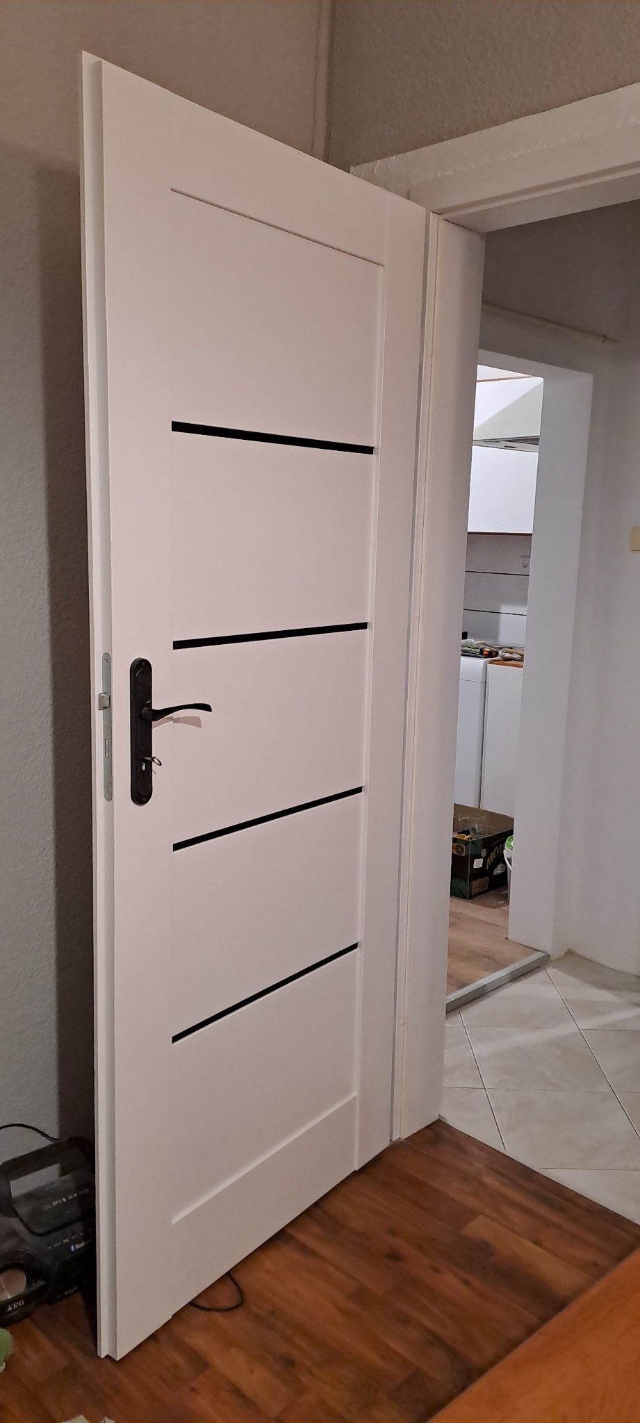 Białe drzwi z czarnymi, poziomymi wstawkami i czarną klamką, otwarte do wnętrza. Widoczna podłoga z paneli i fragment kuchni w tle.