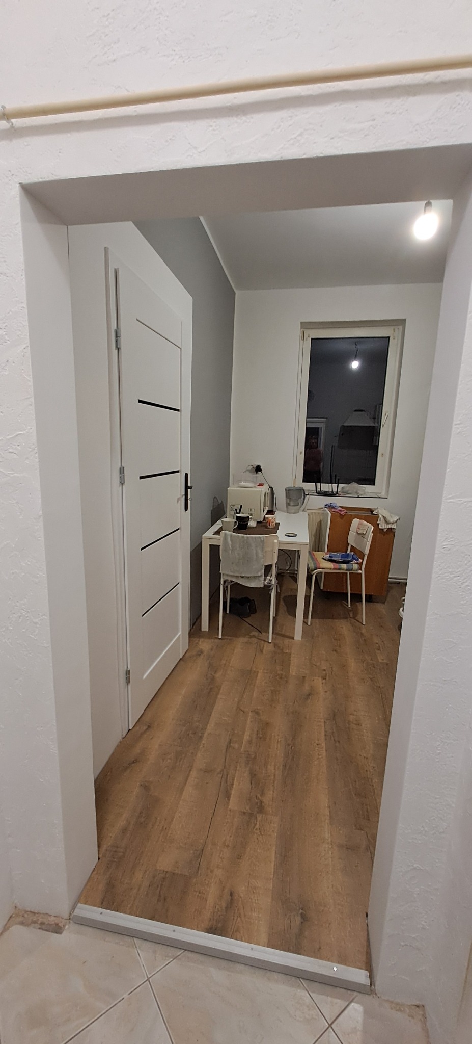 Widok na pokój z drewnianą podłogą przez odnowione, białe futryny i drzwi z czarnymi akcentami. Szara ściana w tle. Widoczne wyposażenie pokoju.