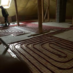 MJ MAGDALENA JOŃCZYK - Instalacja ogrzewania podłogowego na poddaszu: czerwone rury ułożone spiralnie na izolacji, widoczne elementy konstrukcji drewnianej i pracownik w oddali.