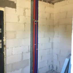 Instalacja wodna z rur w kolorach niebieskim i czerwonym, pionowo poprowadzona w surowym wnętrzu z betonu komórkowego. Szara rura odpływowa.