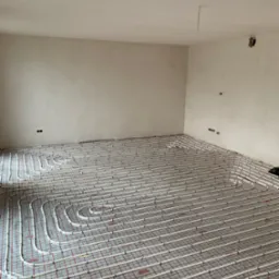 Ułożona instalacja ogrzewania podłogowego z rurami na folii z siatką w surowym pomieszczeniu, widoczne puszki elektryczne w ścianie, gotowe do wylania posadzki.