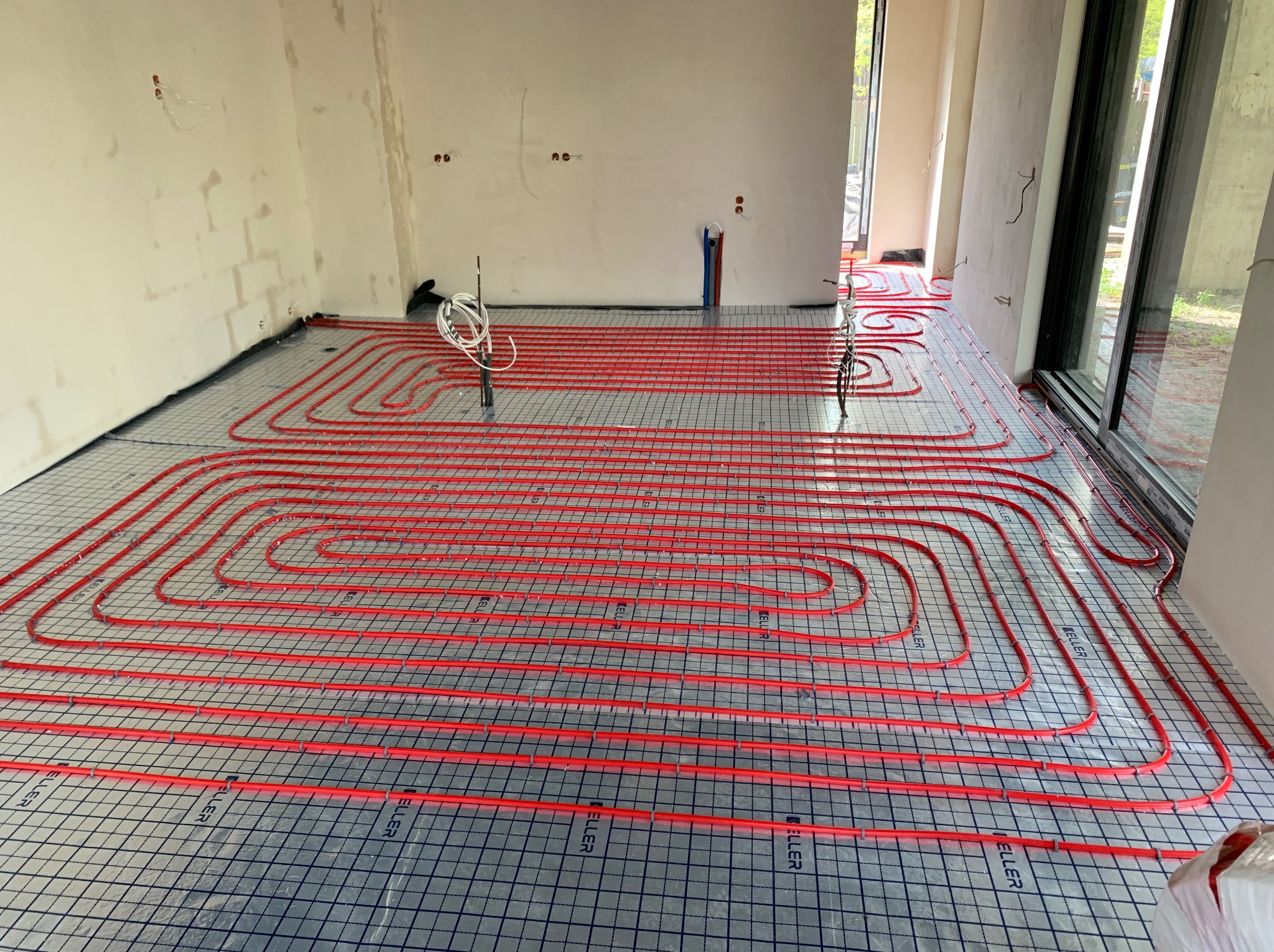 Czerwone rury ogrzewania podłogowego ułożone na srebrnej macie z czarną siatką w nowym budynku w trakcie budowy. Widoczne wystające kable i fragment okna.