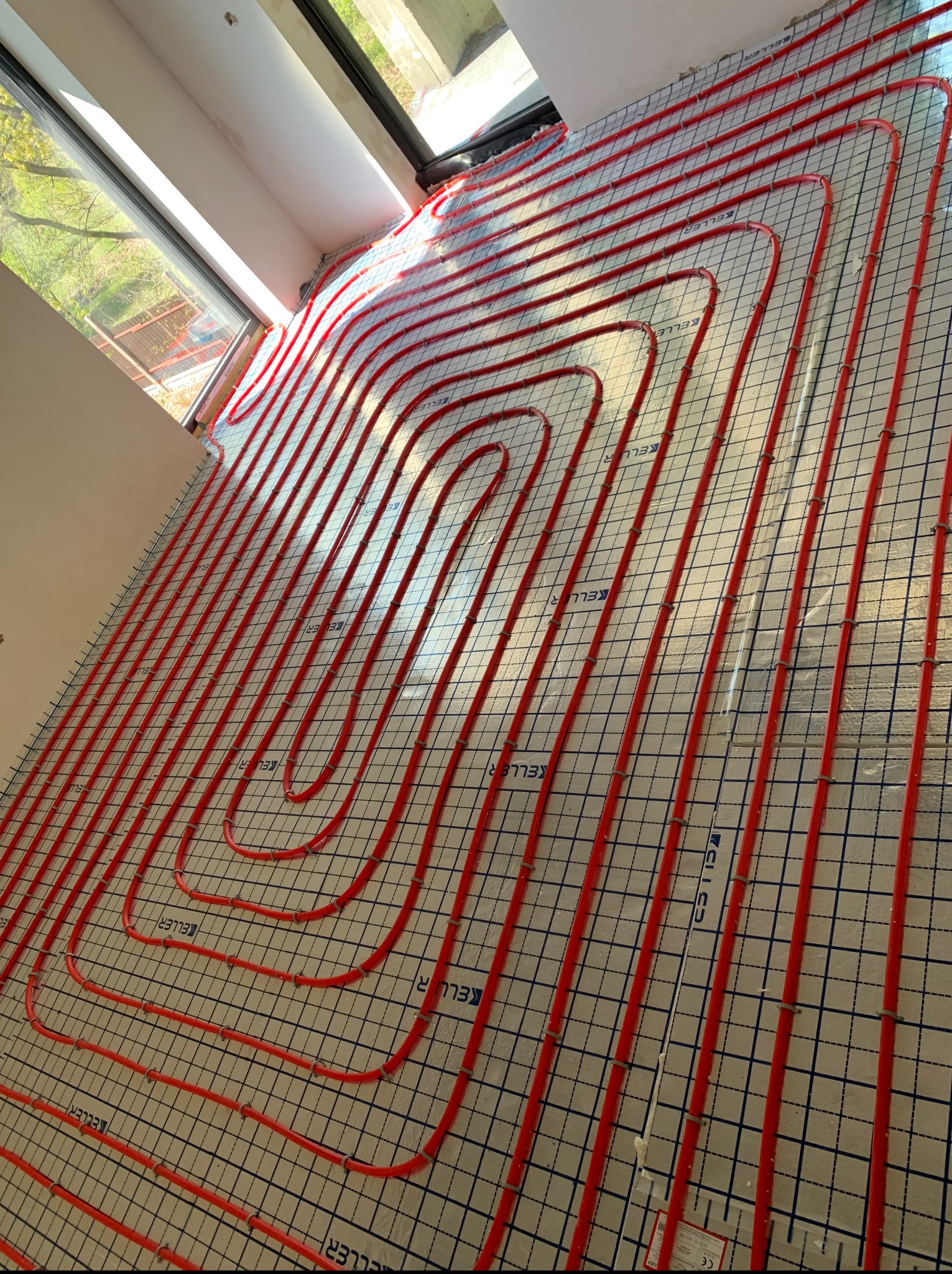 Czerwone rury ogrzewania podłogowego ułożone na izolacji z siatką, widok z góry, w tle okno z widokiem na zieleń. System marki Keller.