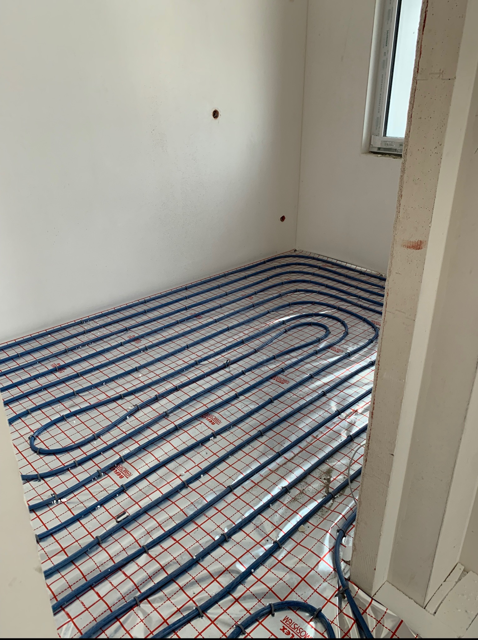 Niebieskie rury ogrzewania podłogowego ułożone na folii z czerwoną kratką w jasnym pomieszczeniu z oknem. Widok z góry, ukośny.