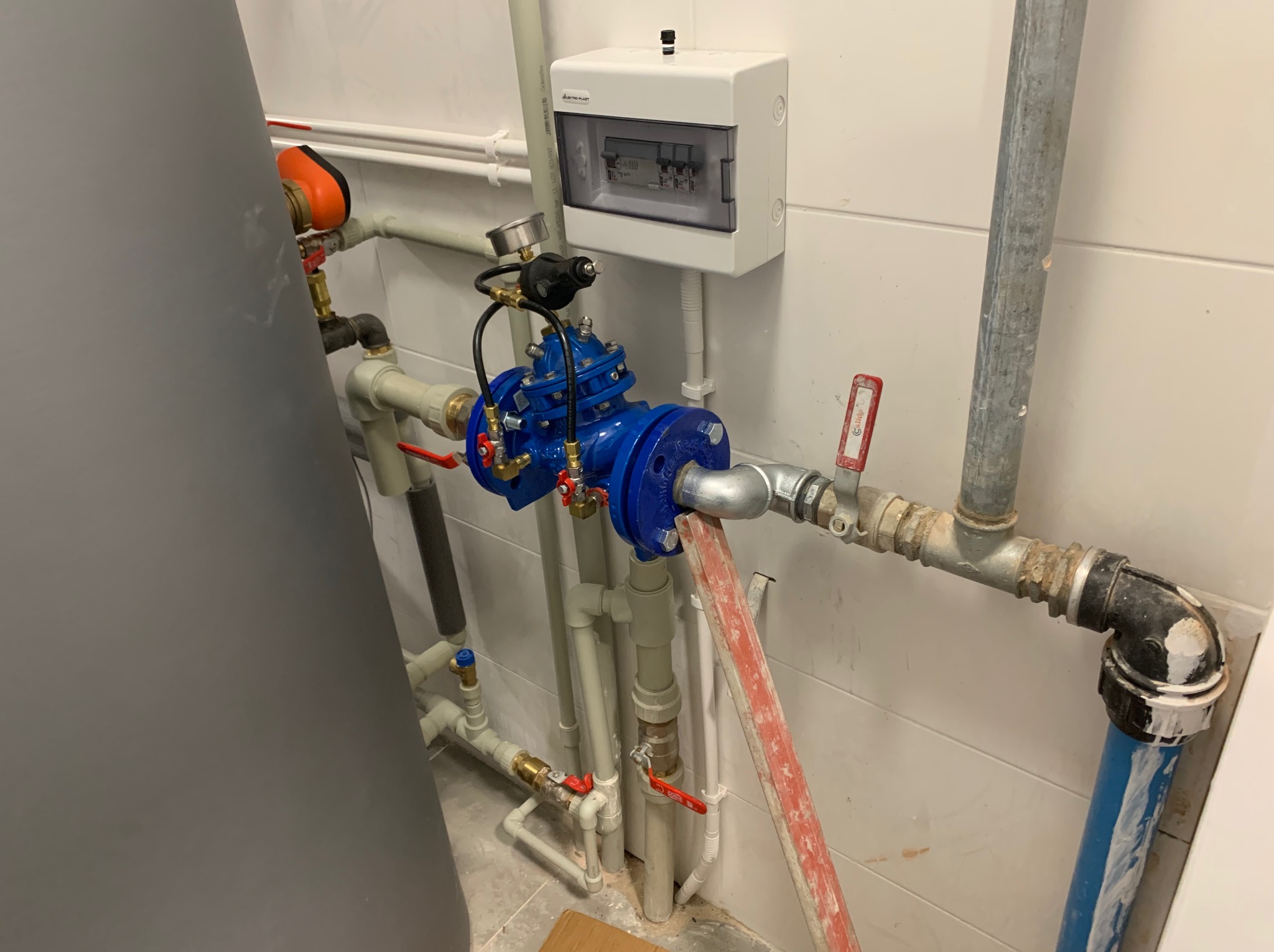 Złożona instalacja hydrauliczna z niebieskim zaworem, manometrem i przewodami. Widoczna skrzynka elektryczna na kafelkowanej ścianie i fragment bojlera.