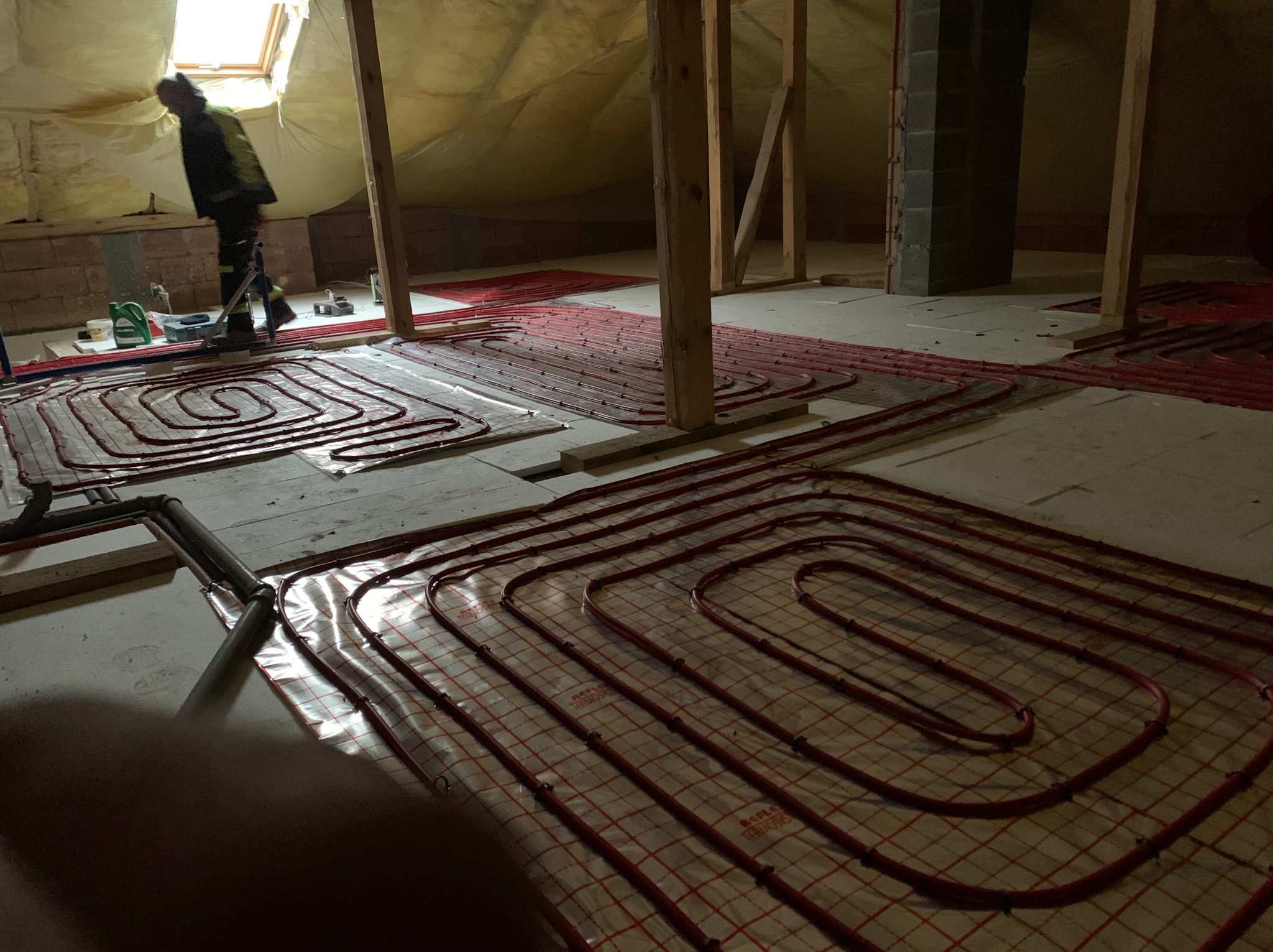 Instalacja ogrzewania podłogowego na poddaszu: czerwone rury ułożone spiralnie na izolacji, widoczne elementy konstrukcji drewnianej i pracownik w oddali.