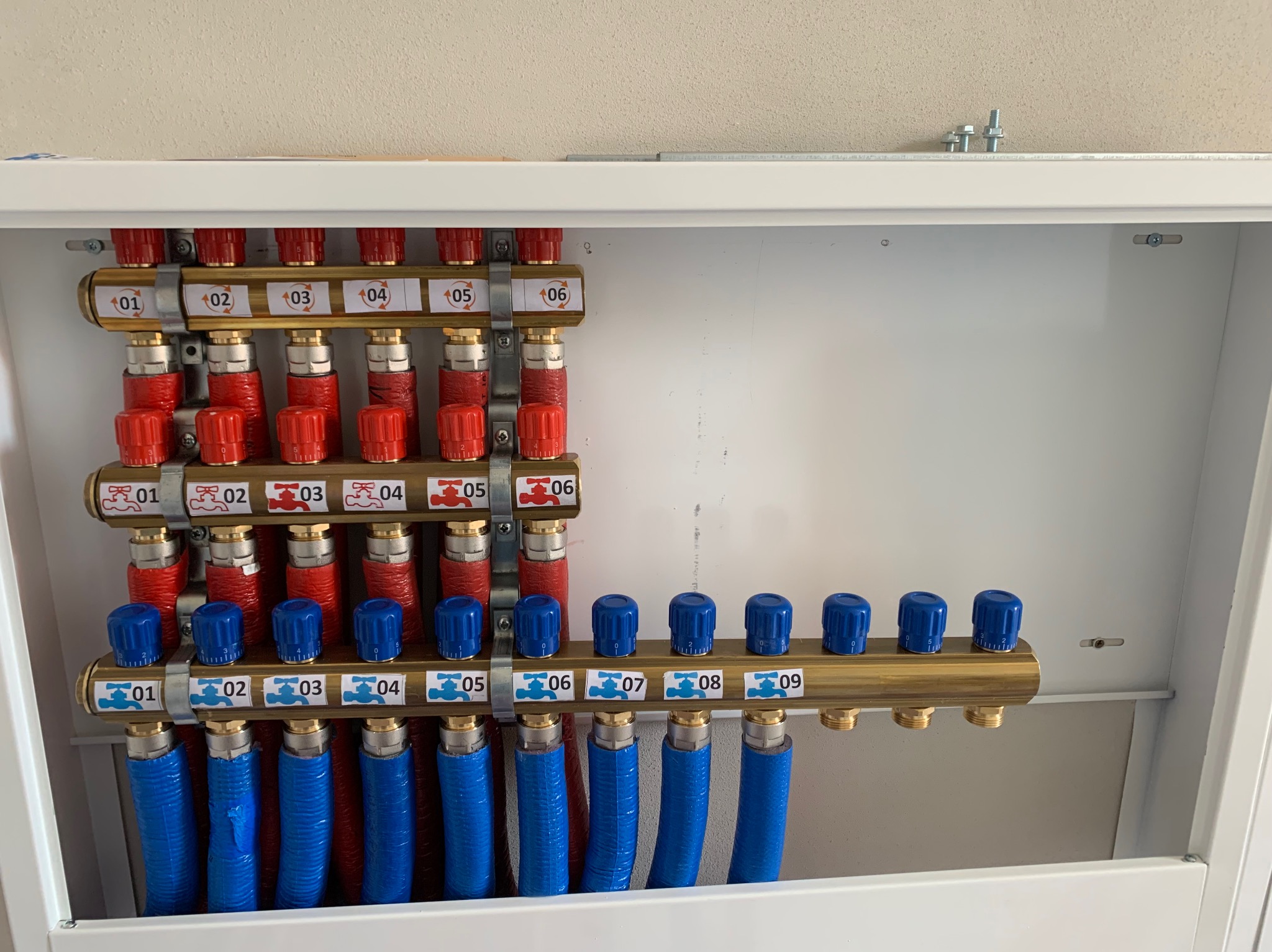 Rozdzielacz ogrzewania podłogowego w szafce, z numerowanymi zaworami i rurami w kolorach czerwonym i niebieskim. Widoczne przyłącza i elementy montażowe.