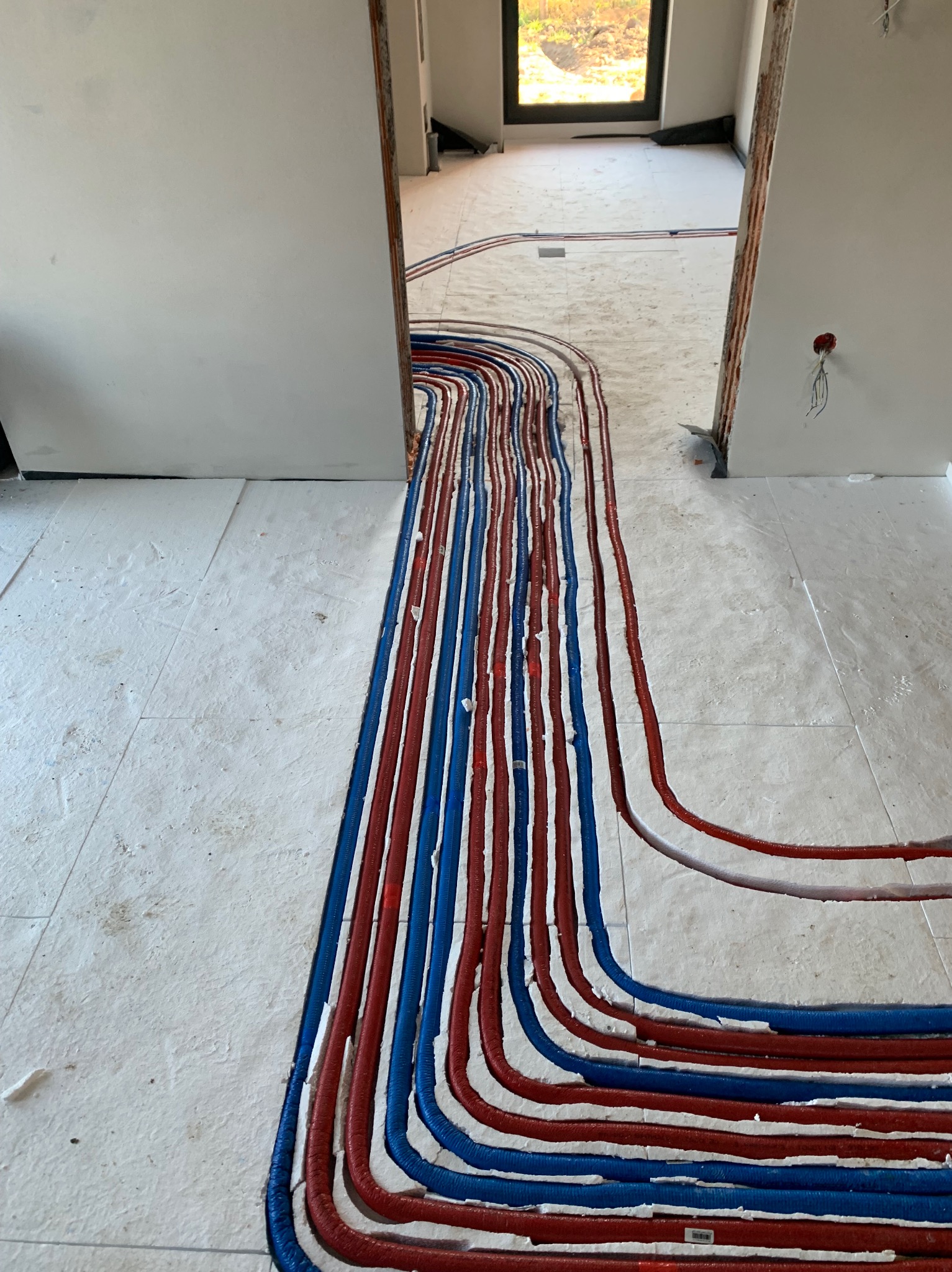 Ułożone, równoległe rury ogrzewania podłogowego w kolorach czerwonym i niebieskim, prowadzące przez próg drzwiowy na jasnej powierzchni podkładu podłogowego.