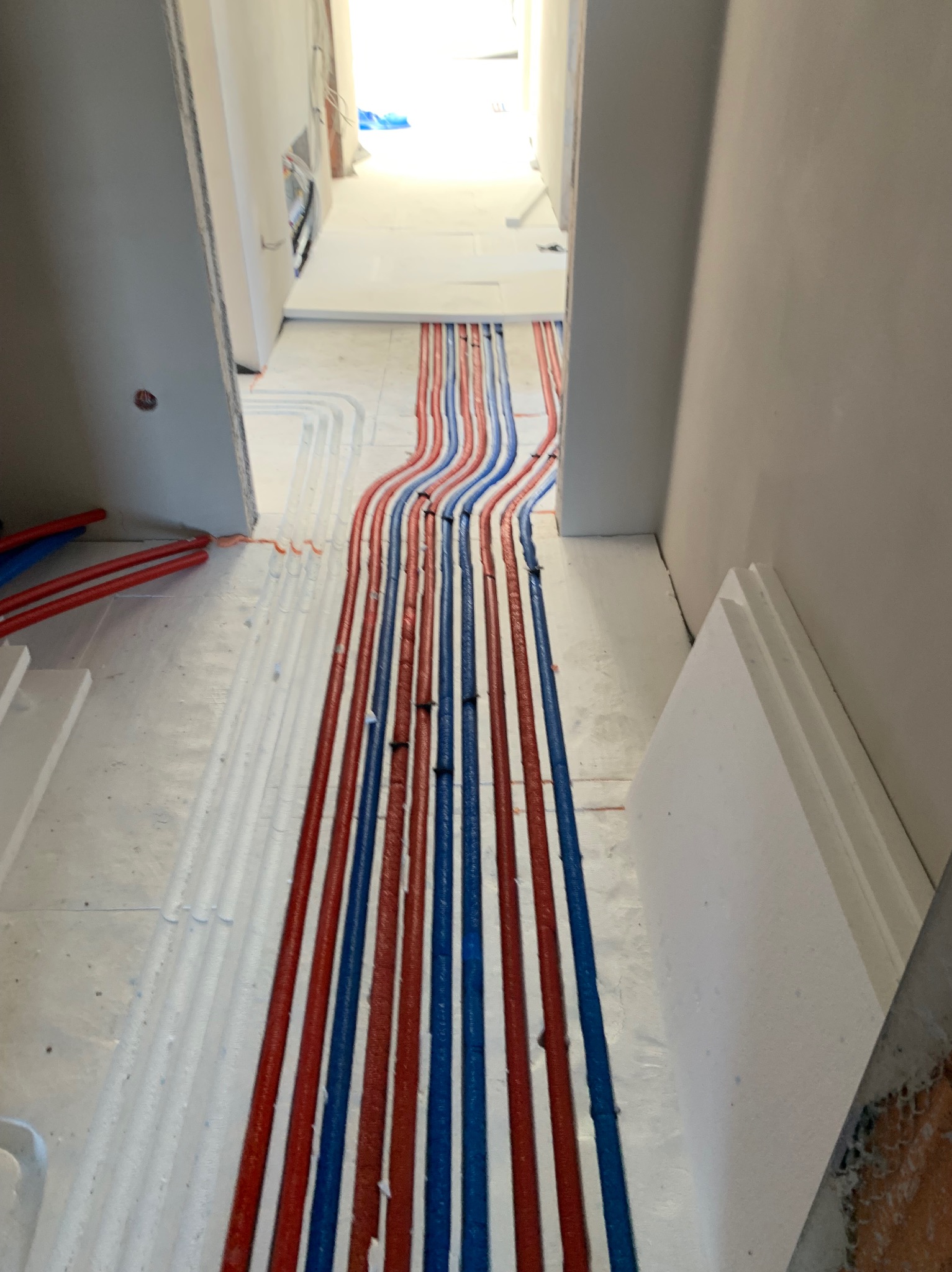 Ułożone, kolorowe rury (czerwone i niebieskie) na białej izolacji podłogowej, przygotowane do zalania posadzką. Widok z perspektywy korytarza w trakcie prac instalacyjnych.
