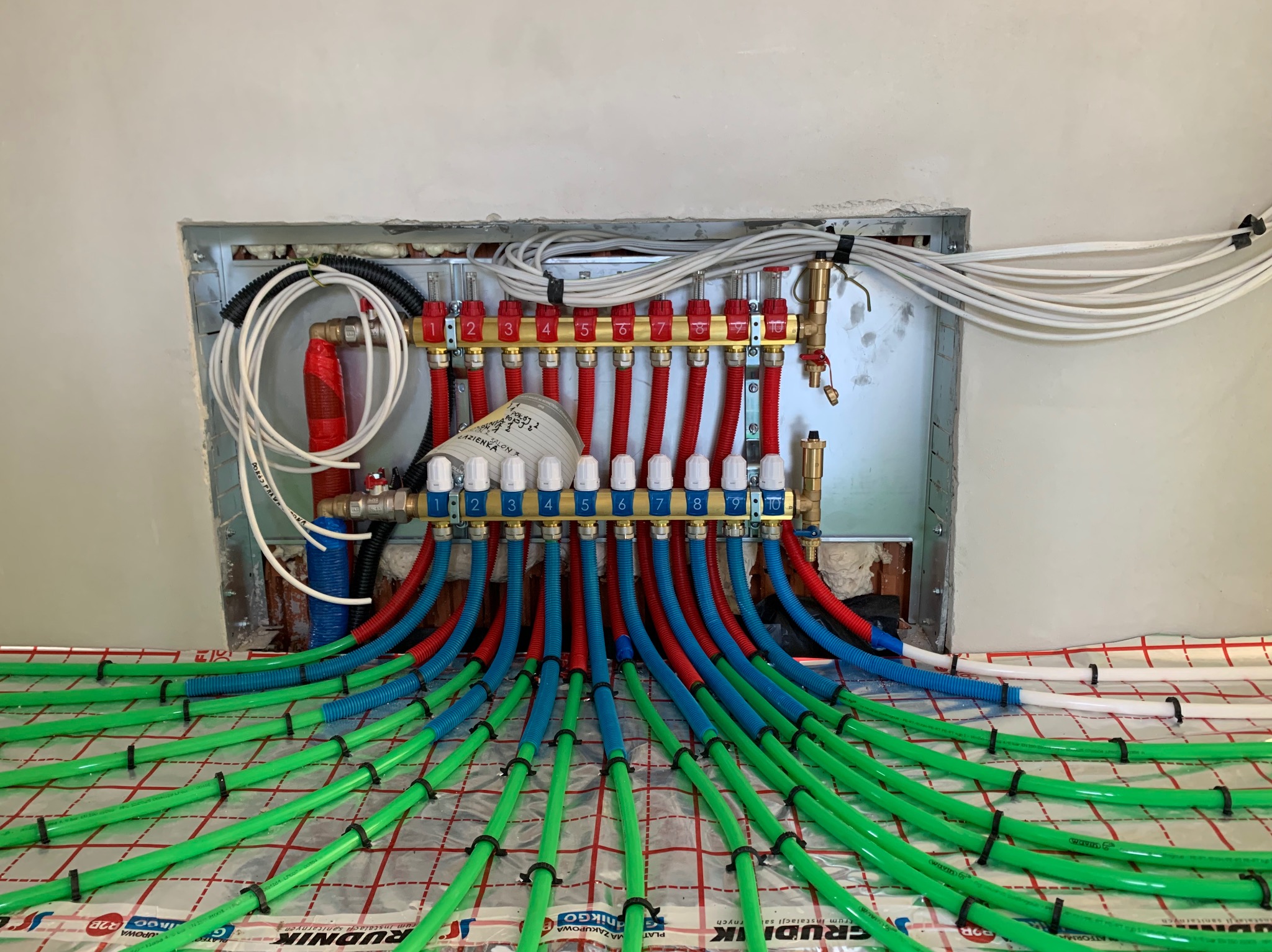 Rozdzielacz ogrzewania podłogowego z rurami w kolorach: zielonym, niebieskim i czerwonym. Widoczne białe przewody i elementy montażowe. Instalacja w trakcie realizacji.