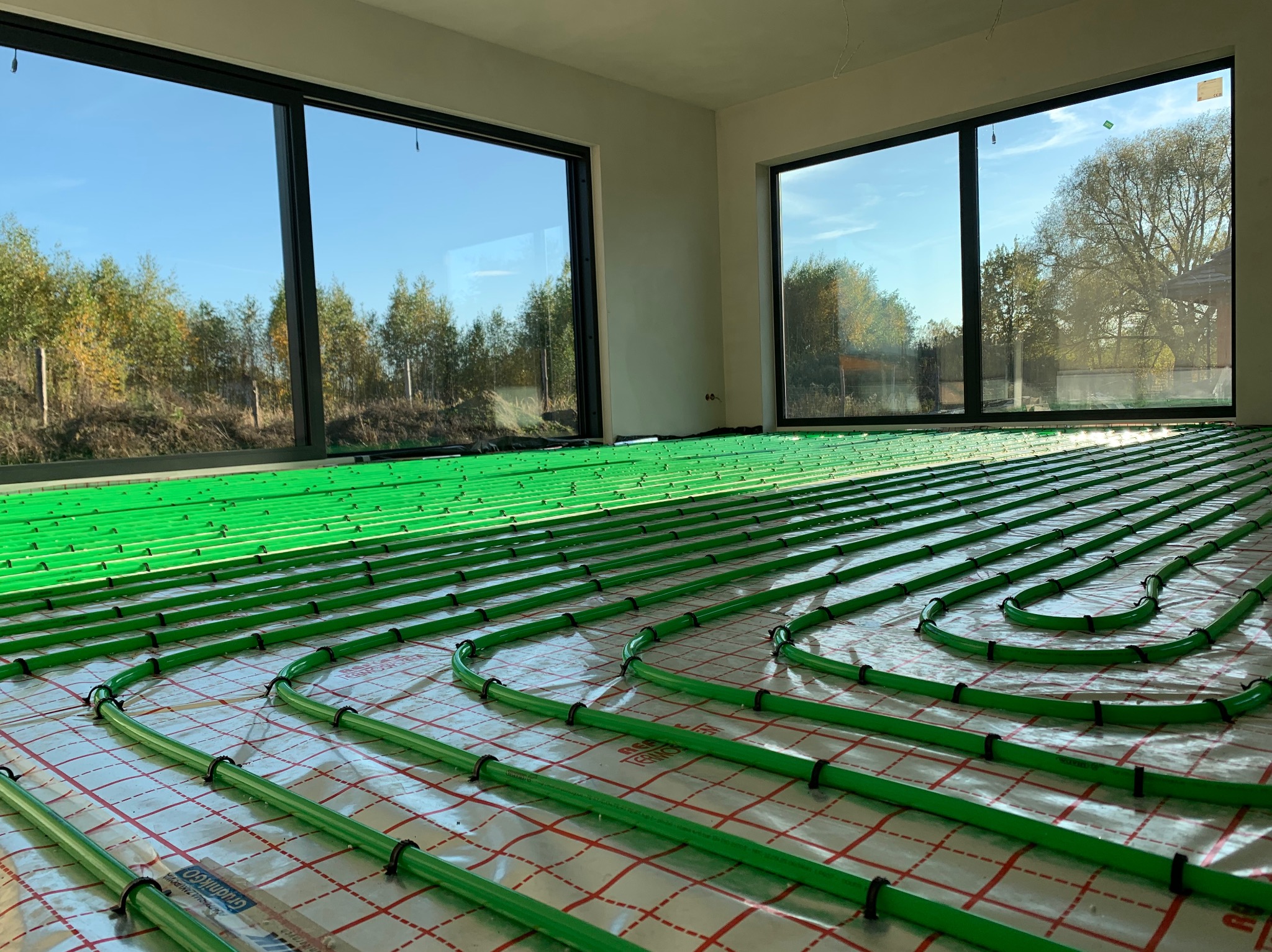 Zielone rury ogrzewania podłogowego ułożone na folii w jasnym pomieszczeniu z dużymi oknami, przez które widać drzewa. Widok z góry, równomierne oświetlenie.