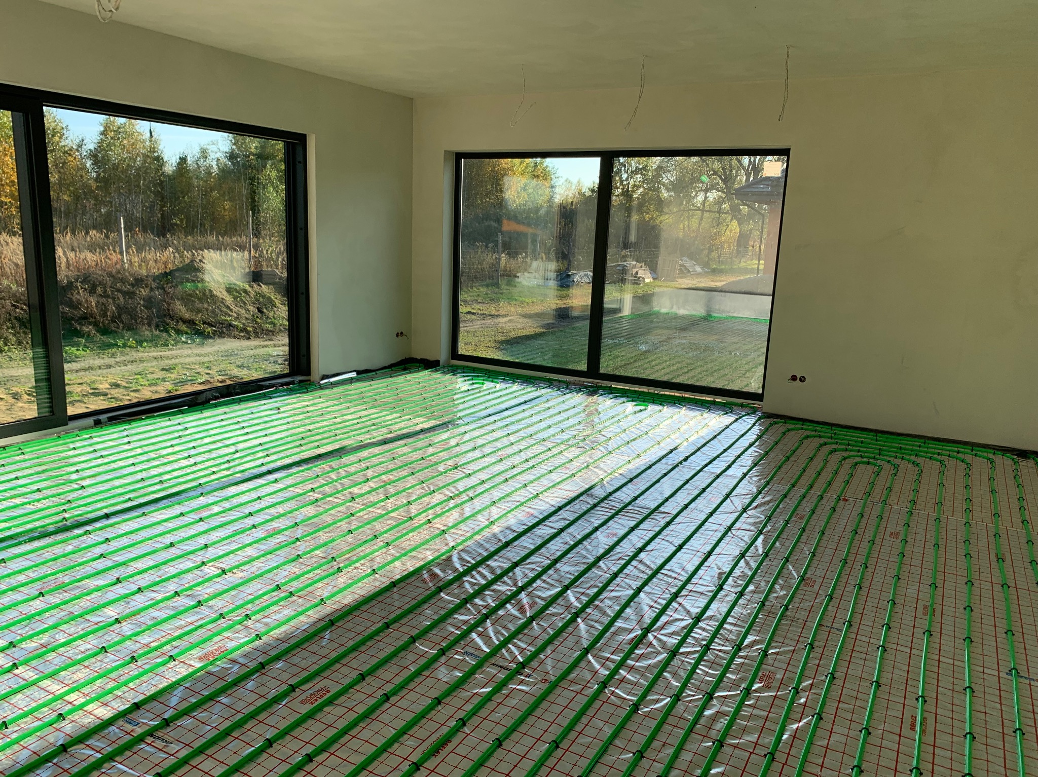 Zielone rury ogrzewania podłogowego rozłożone na folii w jasnym pomieszczeniu z dużymi oknami, widok na zewnątrz. Instalacja przed wylaniem posadzki.