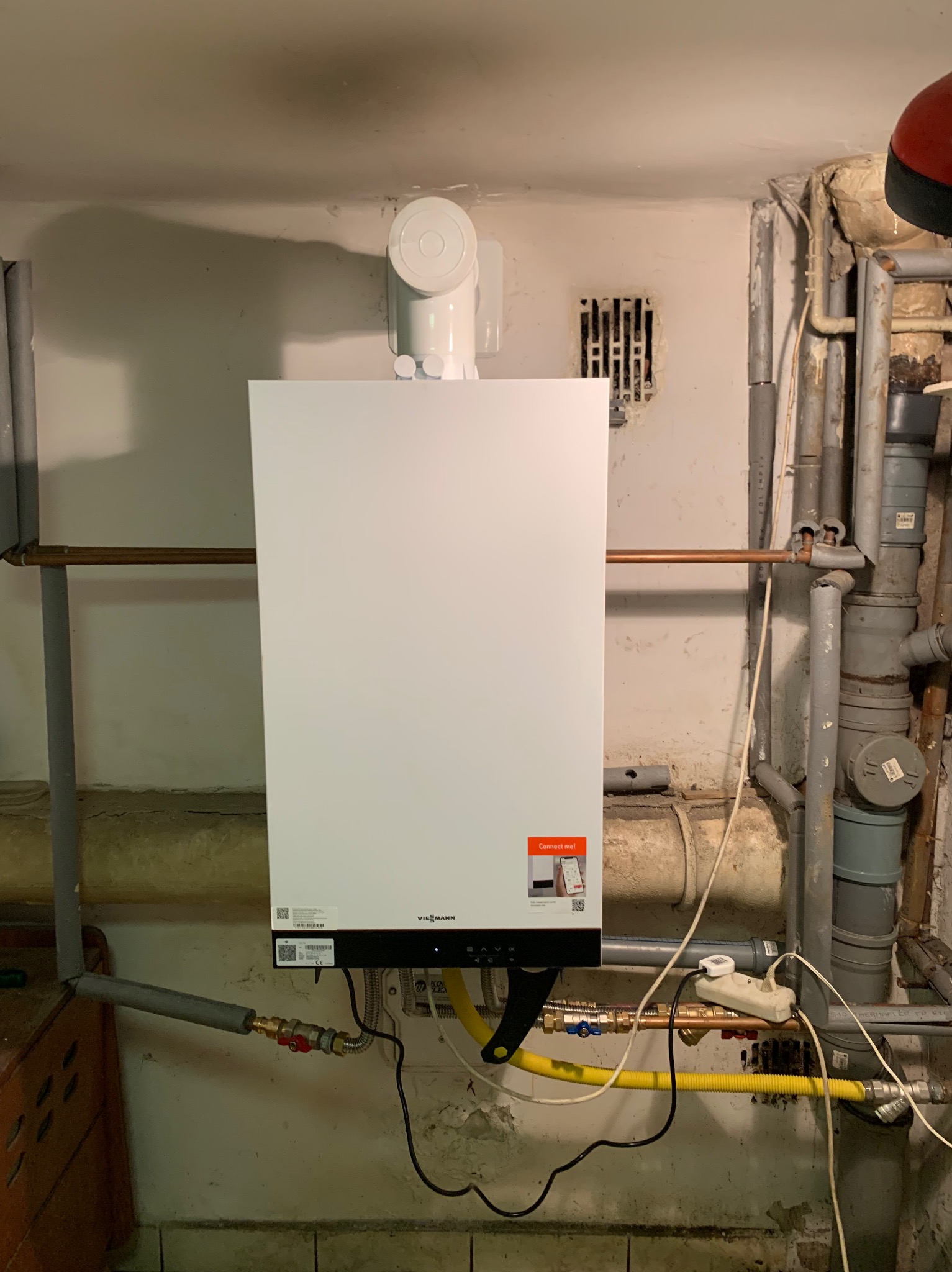 Biały piec gazowy Viessmann zamontowany na ścianie z widocznymi rurami i przewodami. Widoczna kratka wentylacyjna i fragment instalacji.