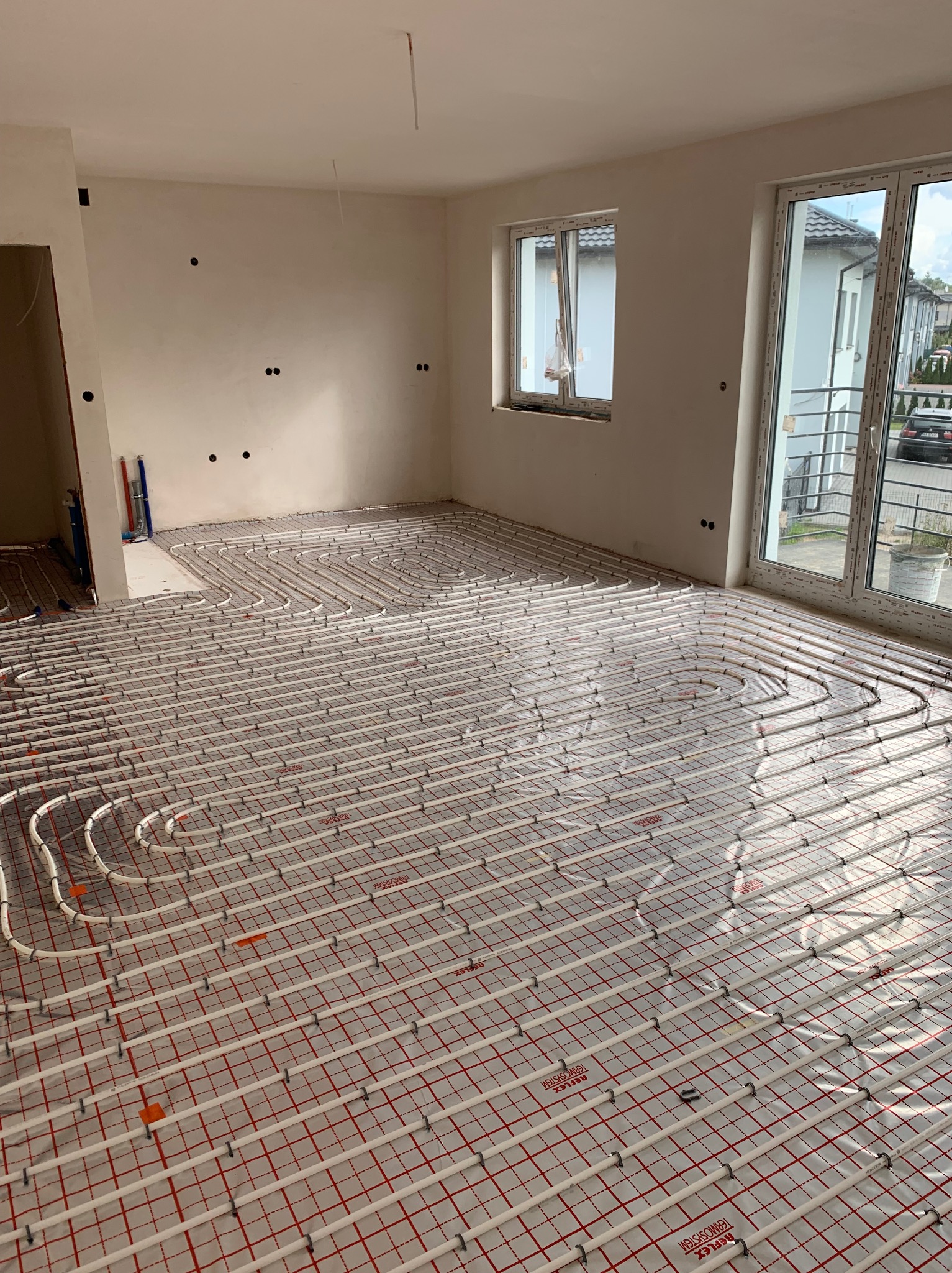 Białe rury ogrzewania podłogowego ułożone na czerwonej siatce w jasnym, surowym wnętrzu z oknami. Widoczne elementy instalacji i przygotowanie do wylewki.