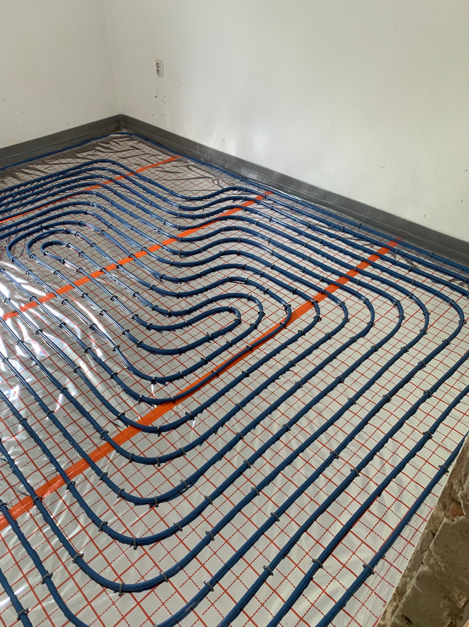 Niebieskie rury ogrzewania podłogowego ułożone na folii z siatką w nowym budynku. Widoczne mocowania rur i fragment ściany z gniazdkiem elektrycznym.