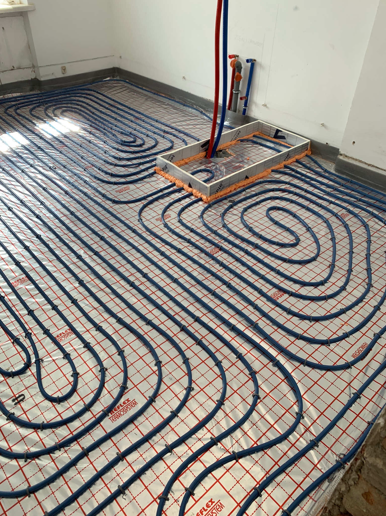 Niebieskie rury ogrzewania podłogowego ułożone na folii z siatką, widoczne pionowe rury doprowadzające wodę w kolorze niebieskim i czerwonym, przygotowane do zalania wylewką.