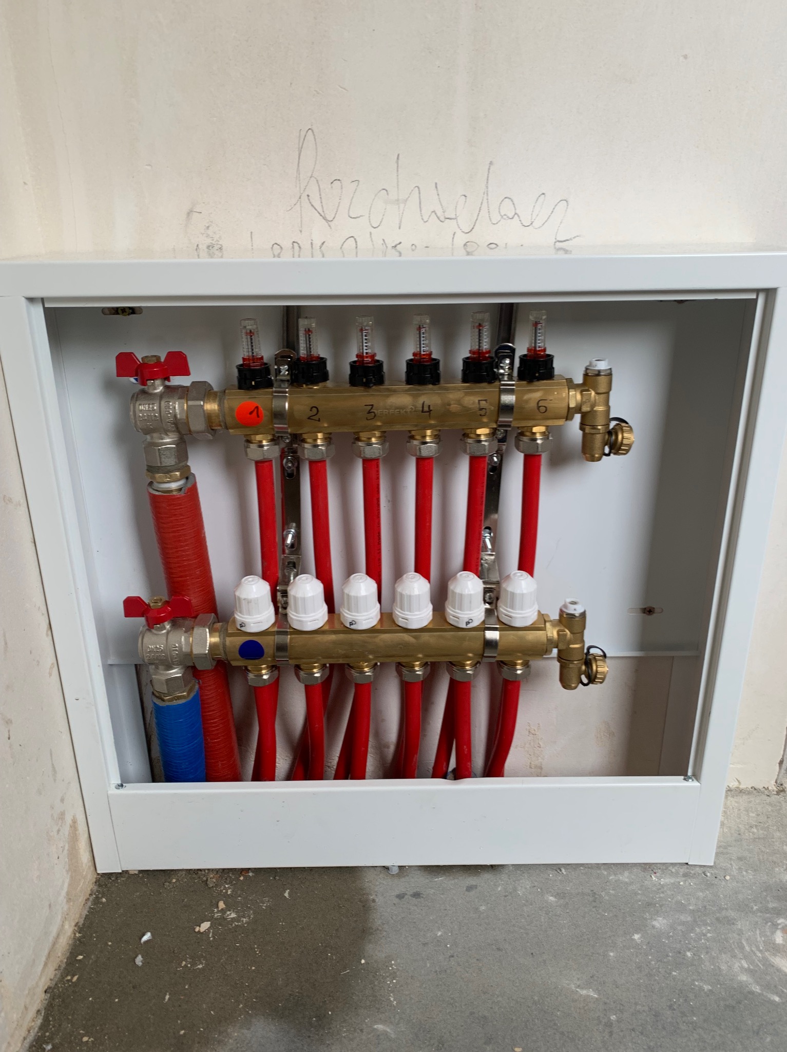 Rozdzielacz ogrzewania podłogowego w białej szafce, z czerwonymi rurami i elementami regulacyjnymi. Widoczne numery na górnej listwie rozdzielacza.