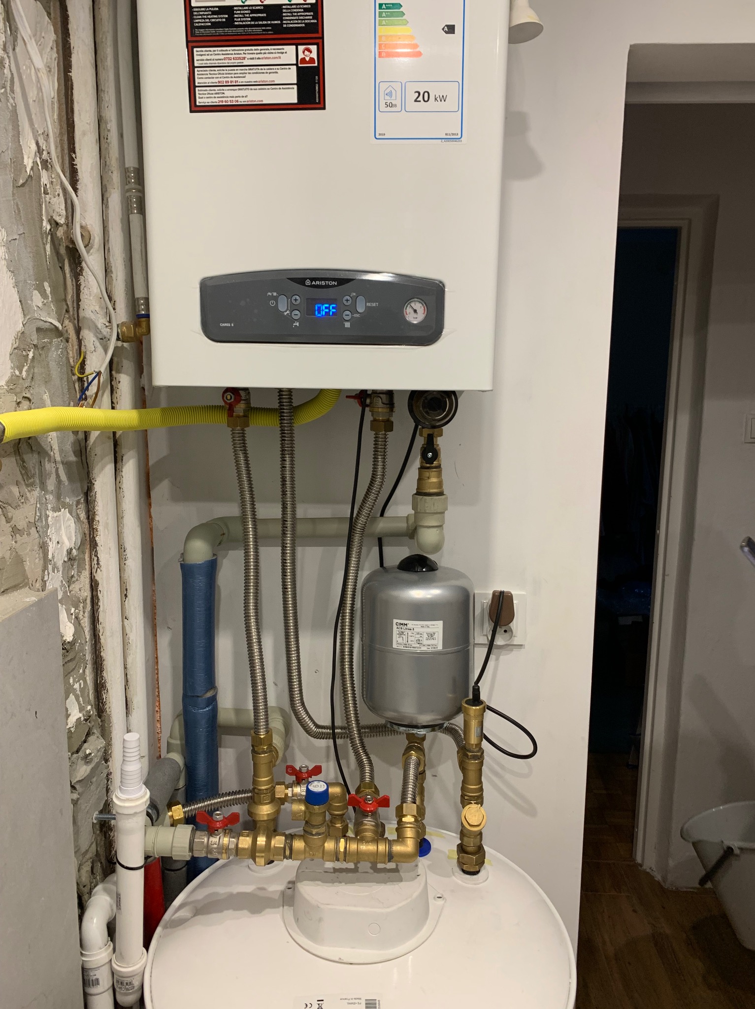 Biały piec gazowy Ariston z szarym panelem sterowania, podłączony do instalacji z miedzianymi rurami i zaworami. Widoczne elementy instalacji grzewczej i gazowej w pomieszczeniu.