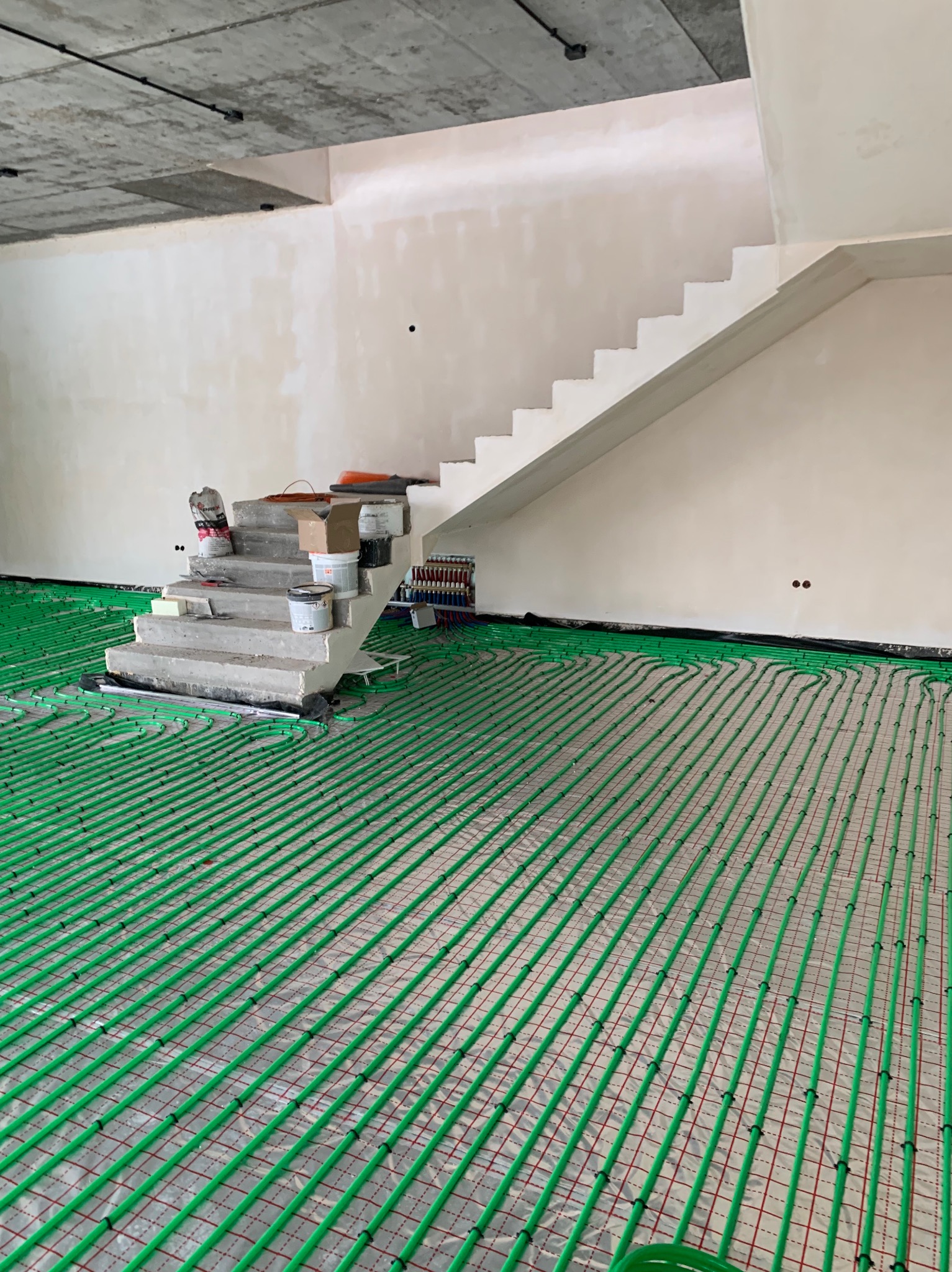 Zielone rury ogrzewania podłogowego ułożone w surowym wnętrzu z betonowymi schodami. Widoczne narzędzia i materiały budowlane. Etap instalacji przed wylaniem posadzki.