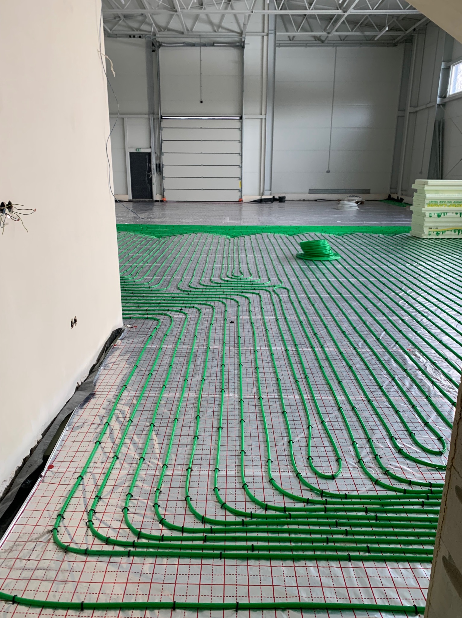 Zielone rury ogrzewania podłogowego ułożone na folii z siatką w dużym, surowym pomieszczeniu. Widoczne elementy konstrukcyjne budynku i materiały izolacyjne.