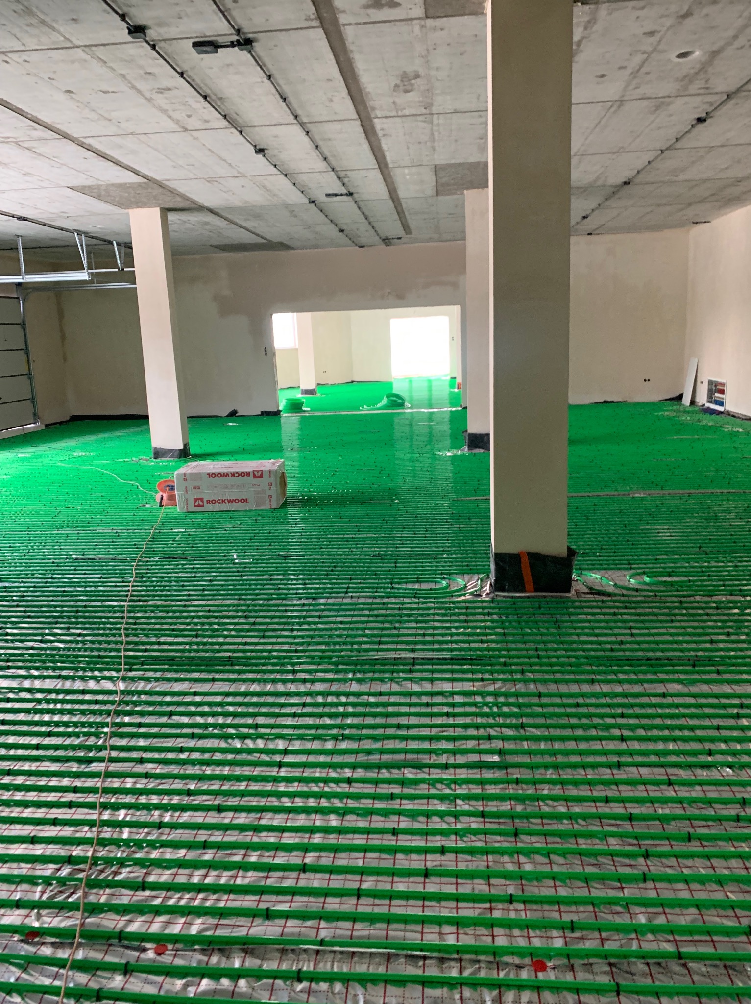 Zielona instalacja ogrzewania podłogowego w surowym wnętrzu, z widoczną izolacją Rockwool. Równoległe rury na folii, przygotowane do zalania posadzką.