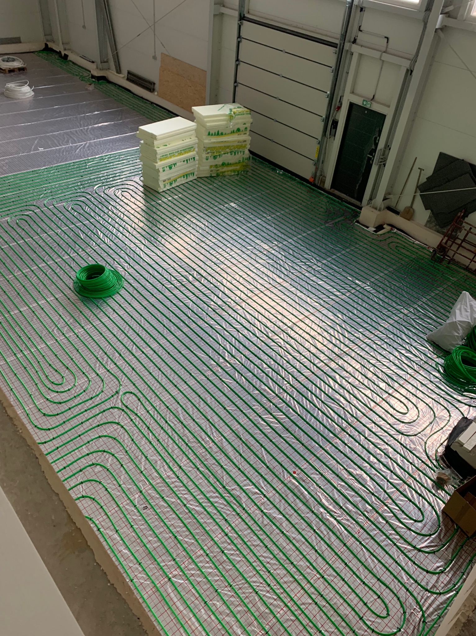 Rozłożona instalacja ogrzewania podłogowego z zielonych rurek na folii, widoczne płyty izolacyjne i zwój rur. Hala z bramą garażową w tle.