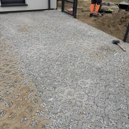 NAJM-BUD - Szara, ażurowa kostka betonowa wypełniona drobnym grysem, ułożona na podjeździe przed domem. Widoczny fragment budynku, furtka i pracownik w odblaskowej odzieży.