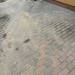 NAJM-BUD - Ułożona kostka brukowa w odcieniach szarości i czerwieni, częściowo posypana piaskiem, przylegająca do ściany budynku. Widoczny fragment osoby i narzędzia.