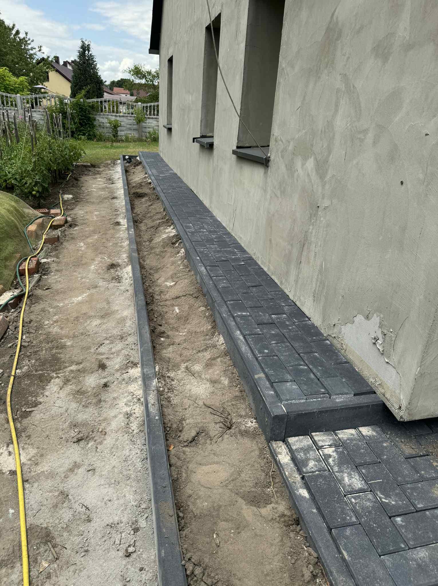 Częściowo ułożona, ciemna kostka brukowa wzdłuż ściany budynku, z widocznym korytkiem drenażowym i przygotowanym podłożem. Prace brukarskie w toku.