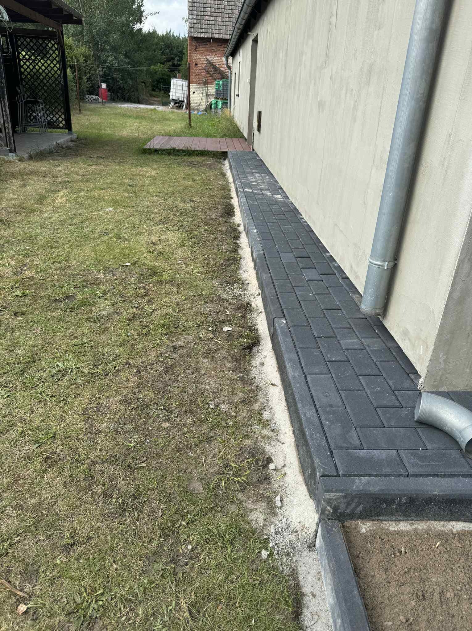 Ułożona, czarna kostka brukowa wzdłuż ściany budynku z rynną, widok z góry. Kostka oddziela trawnik od ściany. Widoczne niedociągnięcia w postaci piasku.