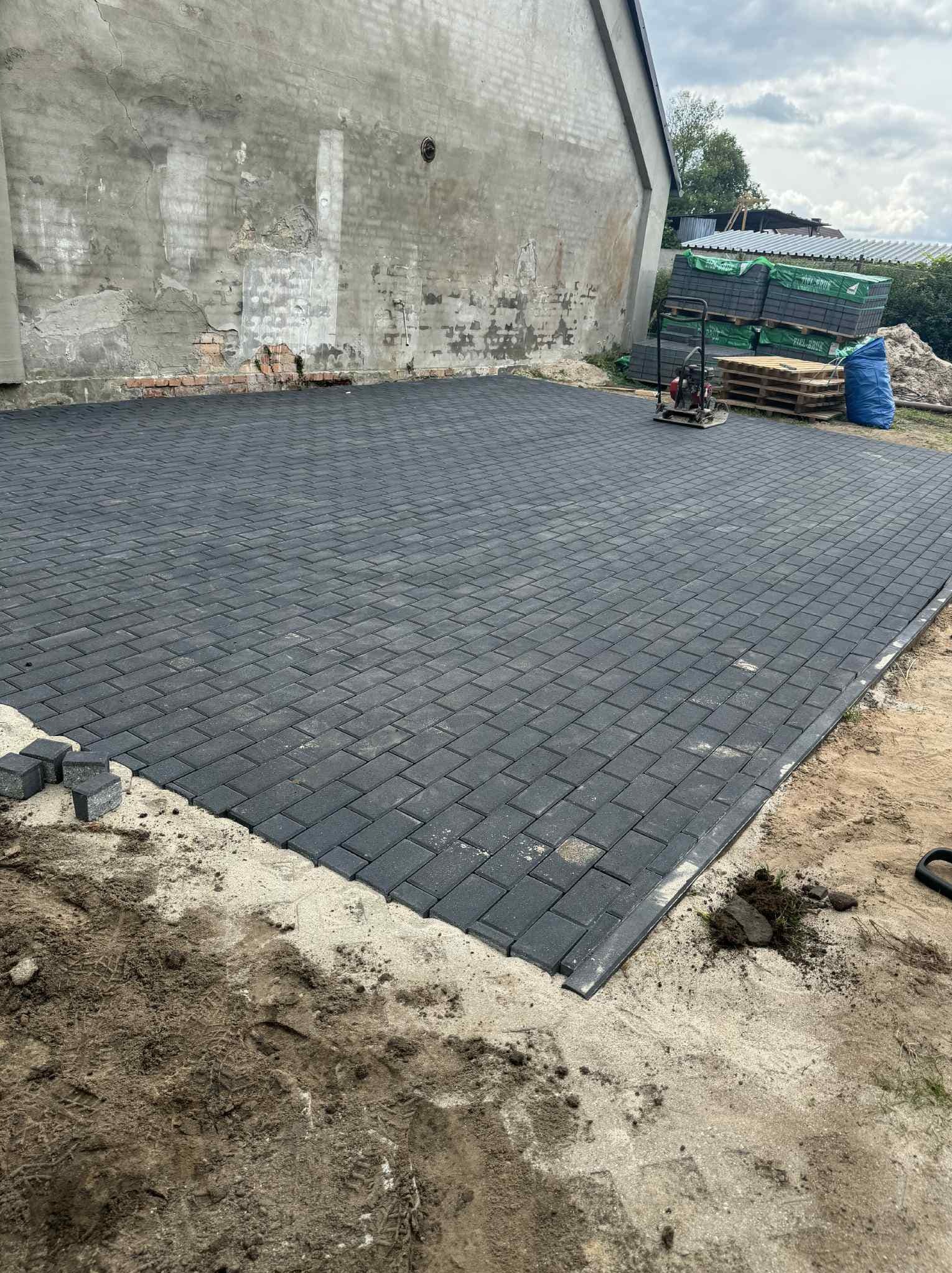 Ułożona, szara kostka brukowa na posesji z widocznym starym budynkiem w tle. Na obrzeżach piasek i ziemia, w rogu paleta z kostką i ubijarka.