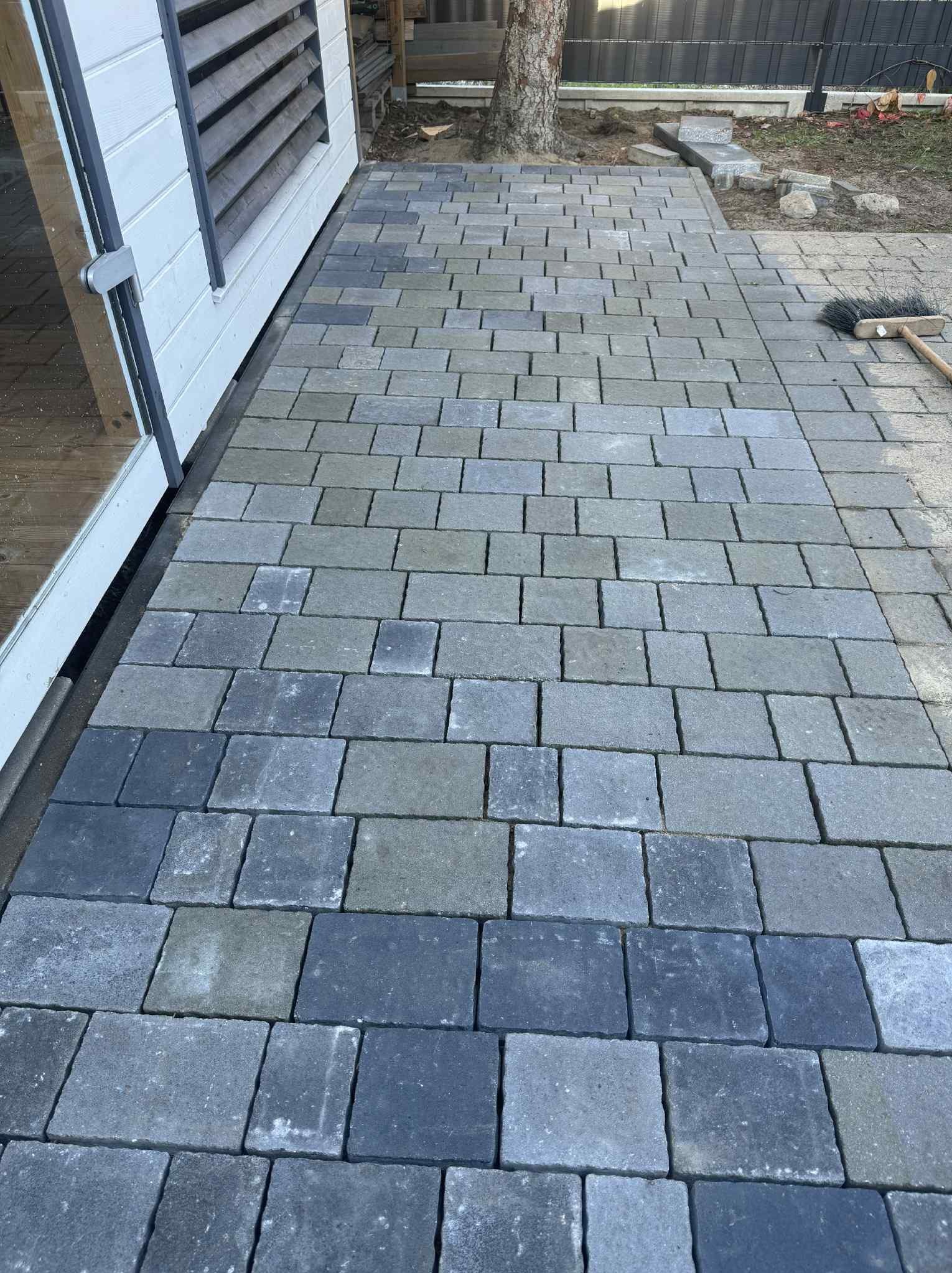 Ścieżka z szarej kostki brukowej prowadząca wzdłuż białej ściany budynku, z widocznym fragmentem drzewa i narzędziem do zamiatania na obrzeżach.
