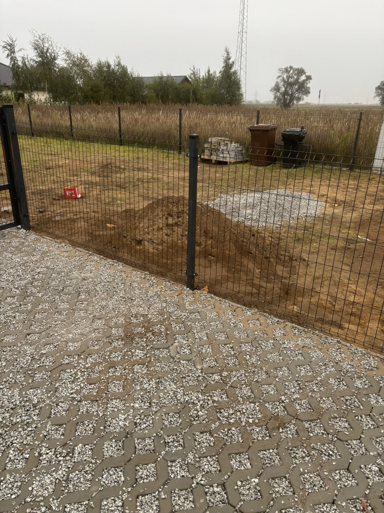 Układanie ażurowej kostki brukowej z wypełnieniem żwirowym przy ogrodzeniu z siatki. Widoczny fragment terenu z ziemią i przygotowanymi materiałami budowlanymi.