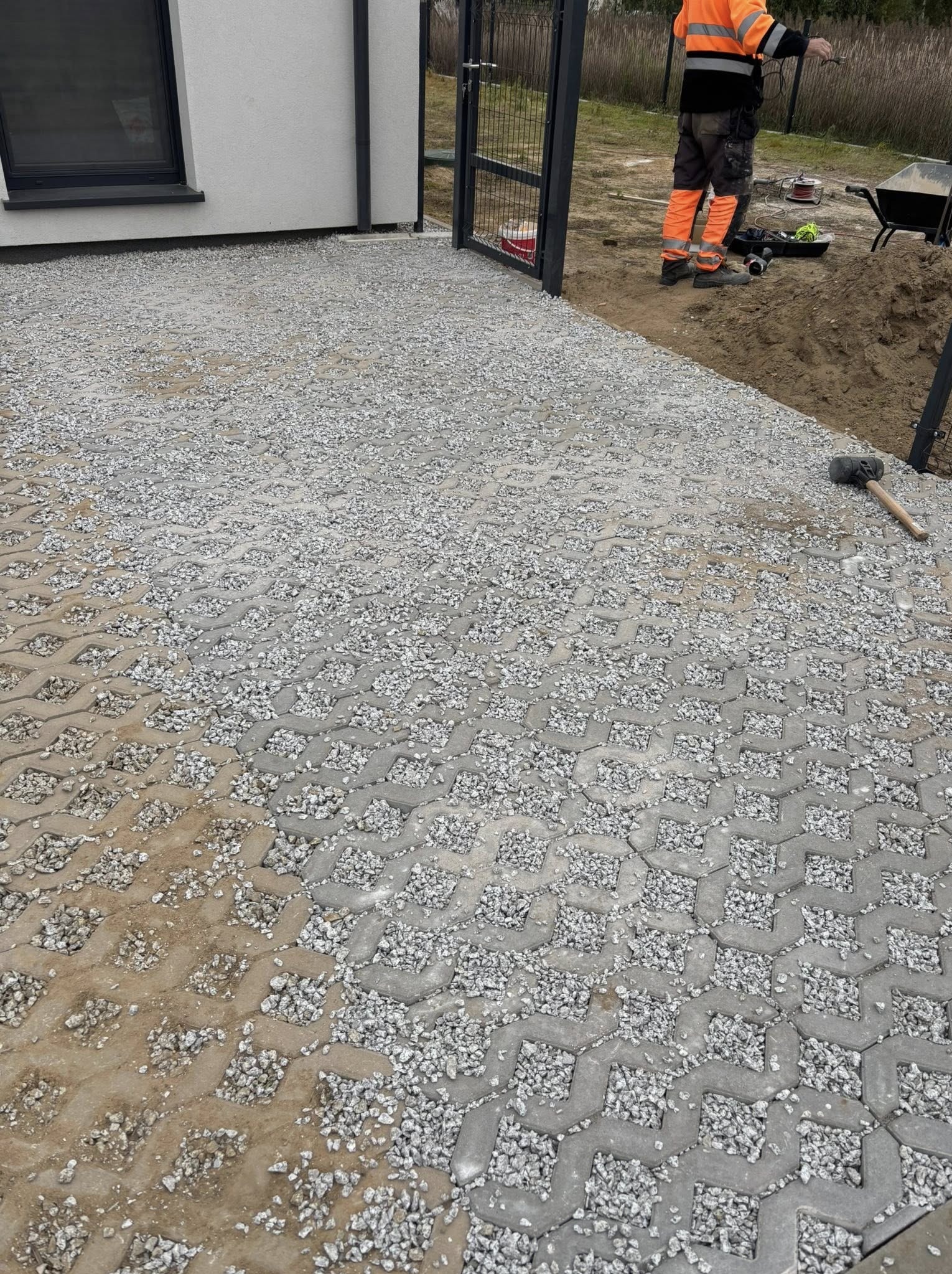 Szara, ażurowa kostka betonowa wypełniona drobnym grysem, ułożona na podjeździe przed domem. Widoczny fragment budynku, furtka i pracownik w odblaskowej odzieży.