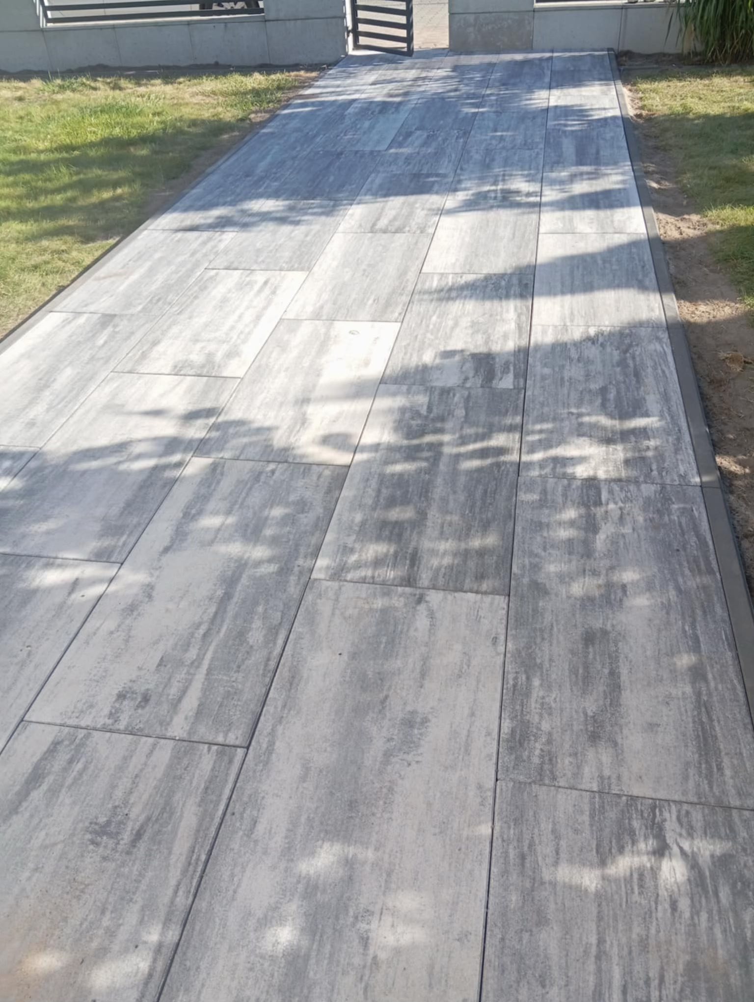 Ścieżka z prostokątnej, szarej kostki brukowej z delikatnym wzorem, obrzeża z ciemnej kostki, trawnik po bokach, brama w tle, cień drzew na powierzchni.