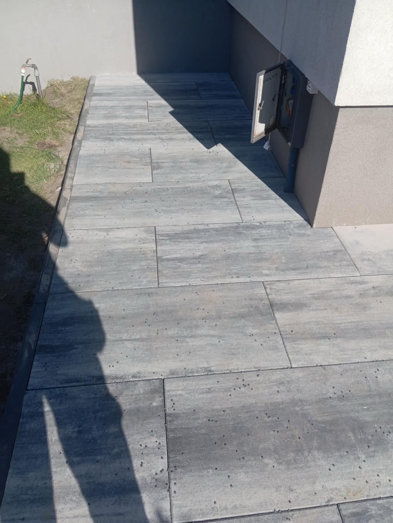 Ścieżka z szarej kostki brukowej prowadząca wzdłuż budynku, z widoczną studzienką rewizyjną i fragmentem trawnika z kranem. Ułożenie poziome, prostokątne.
