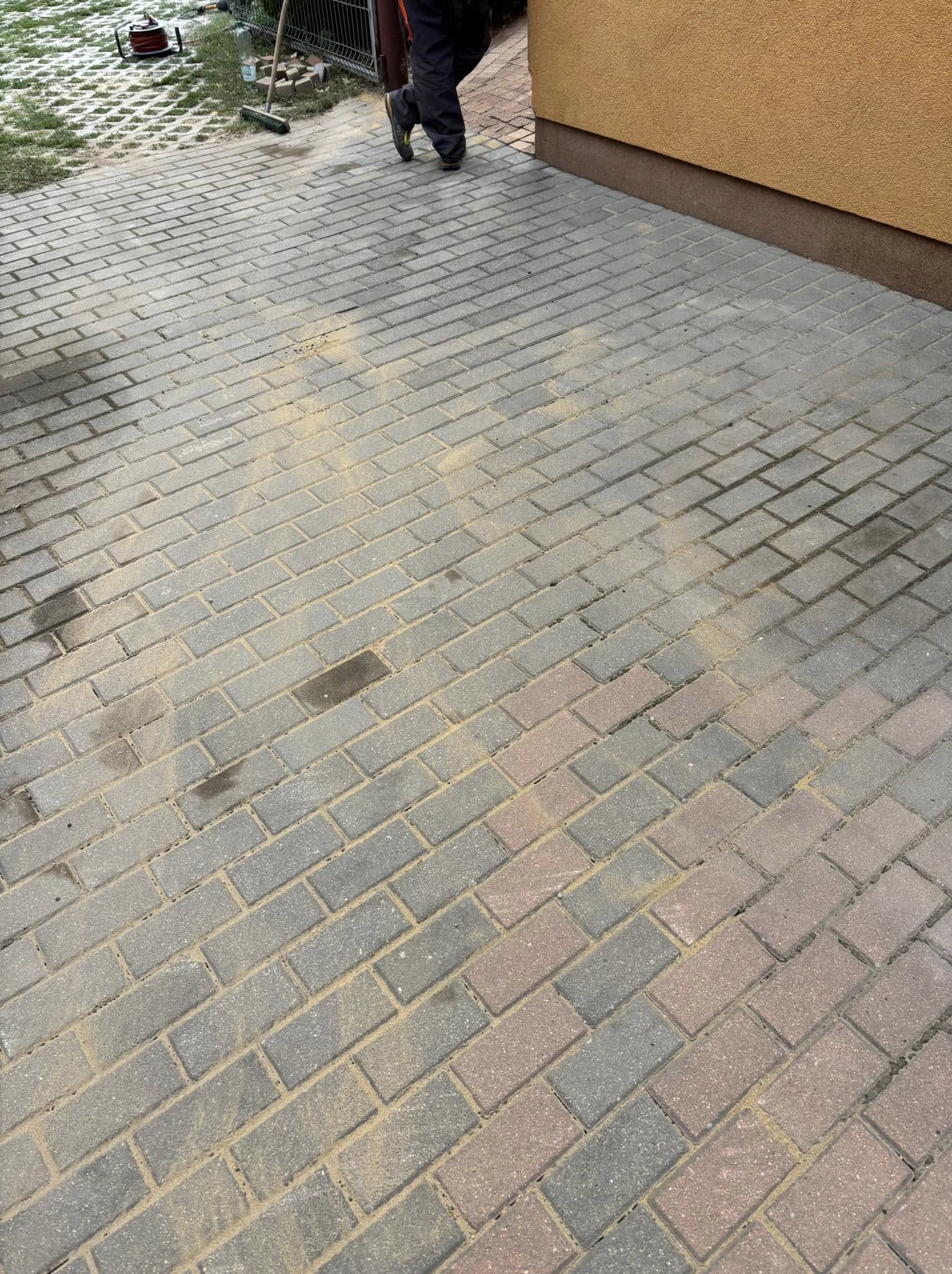 Ułożona kostka brukowa w odcieniach szarości i czerwieni, częściowo posypana piaskiem, przylegająca do ściany budynku. Widoczny fragment osoby i narzędzia.